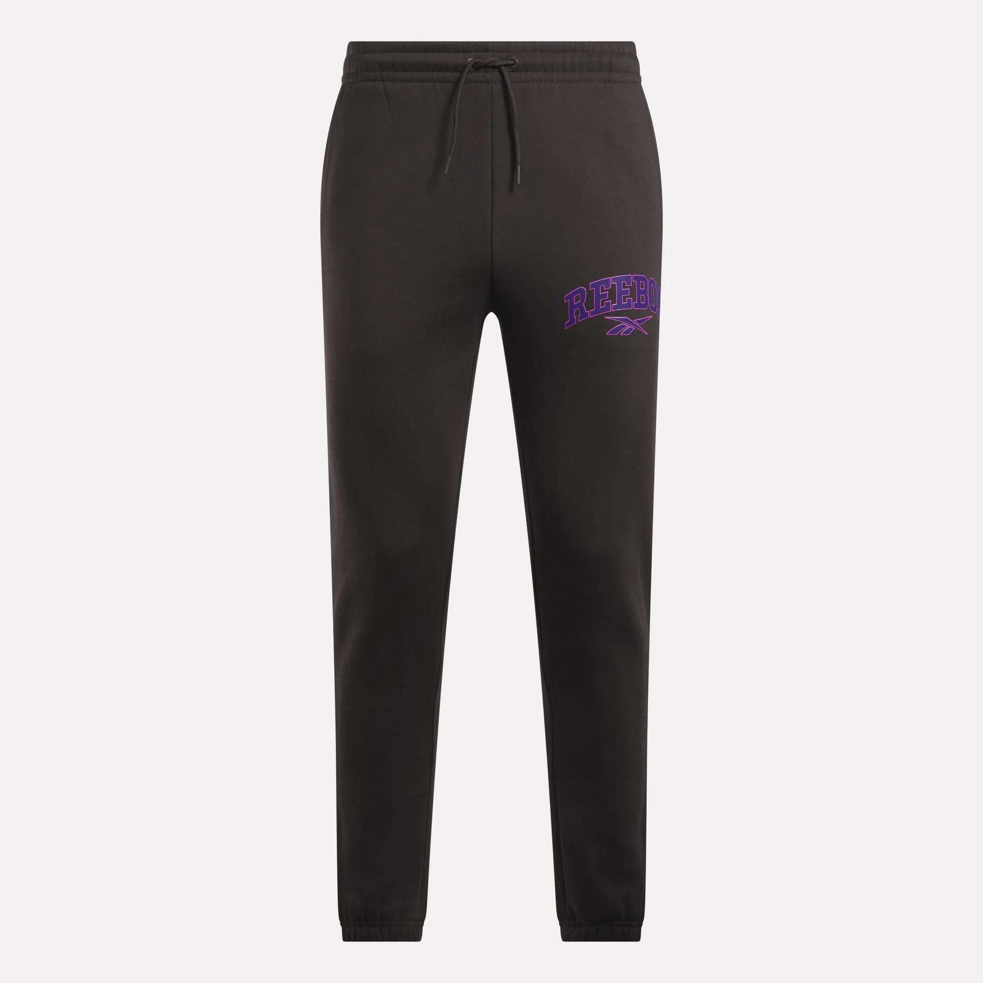 Men's Varsity Joggers by Reebok in black, with a relaxed fit, elastic waistband, drawstring, and cuffed ankles. Features purple REEBOK text and logo on the left thigh. Set against a light gray background.