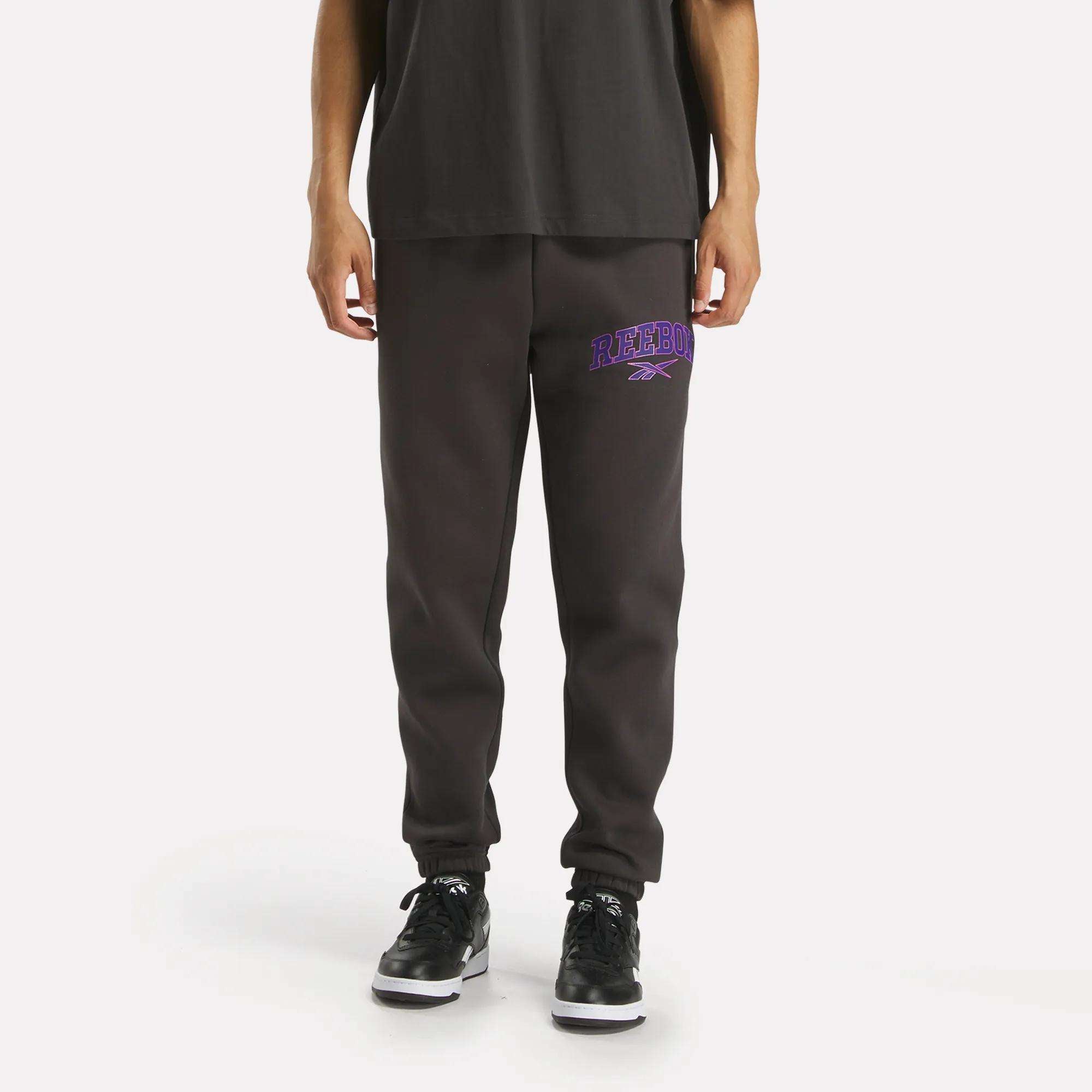 A man in Reebok Men's Varsity Joggers, dark gray with a purple Reebok logo on the left thigh, pairs them with a black shirt and sneakers while standing against a white background to highlight their relaxed fit.