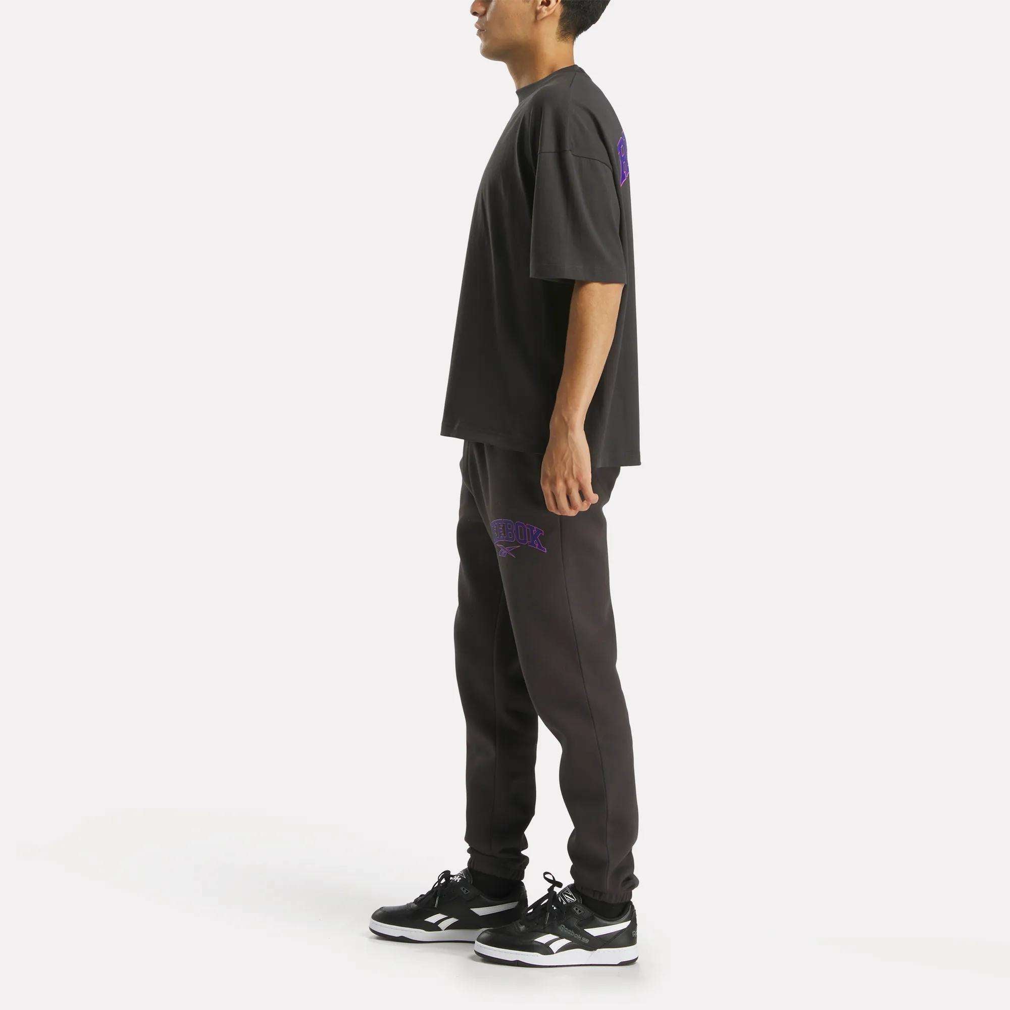 A person stands sideways in a relaxed fit black T-shirt, matching Reebok Men's Varsity Joggers, and black-and-white sneakers against a plain white background.