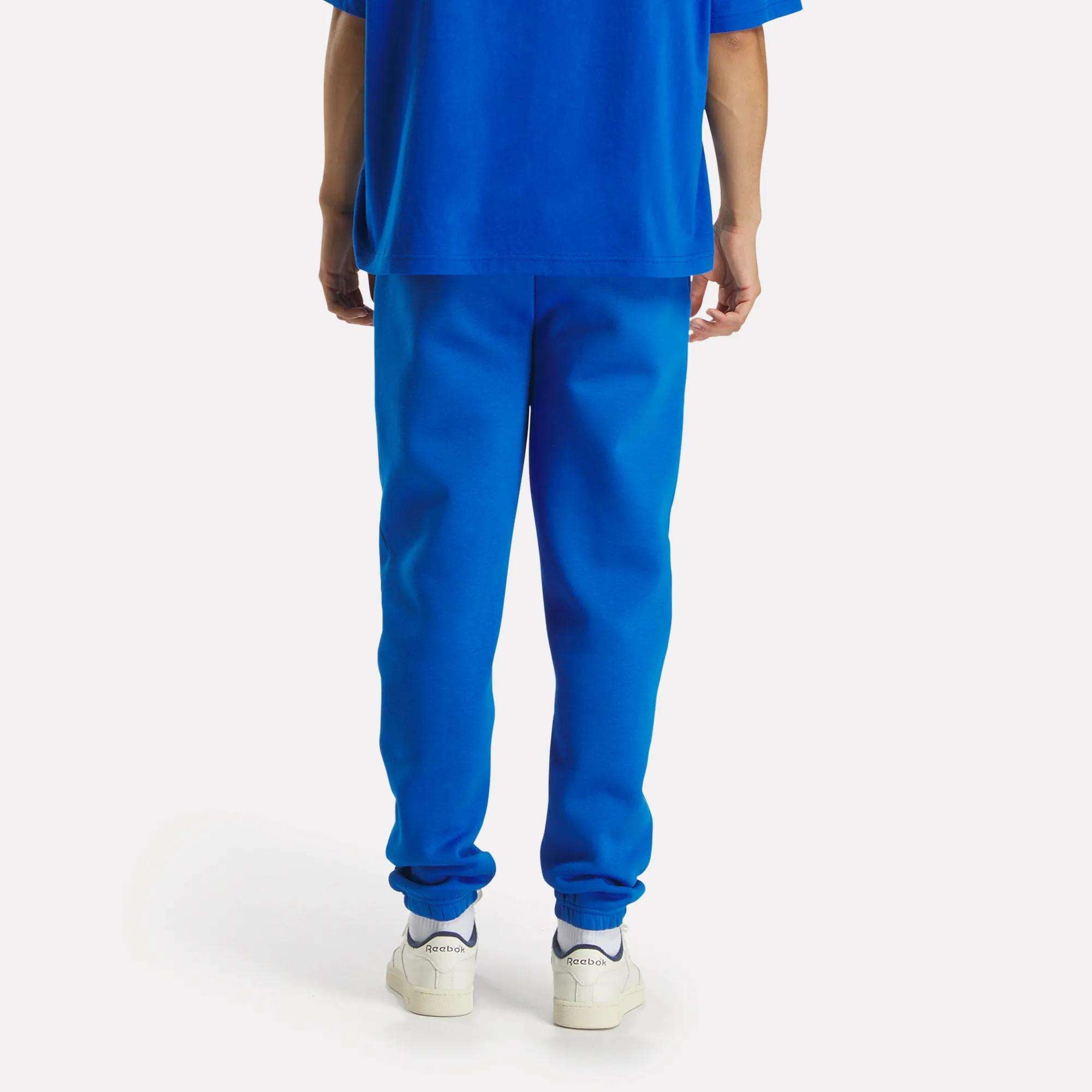 A person stands facing away, wearing Reebok Men's Varsity Joggers in a relaxed fit with a bright blue tee. White Reebok sneakers with visible branding on the heels complete the look against a plain white background.