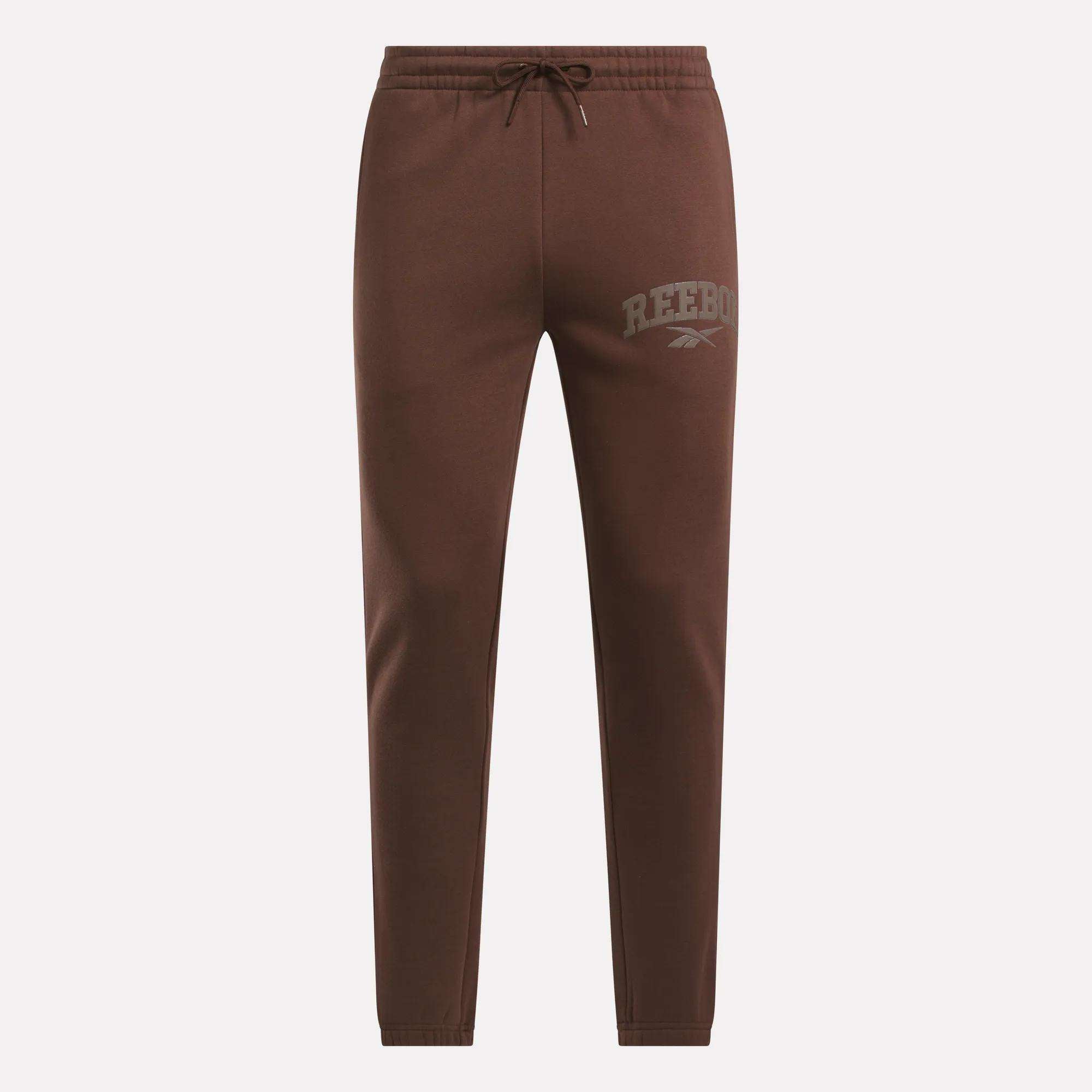 Reebok Men's Varsity Joggers in brown feature an elastic waistband, drawstring, and Reebok logo on the left thigh. These slim-fit joggers have no visible pockets, combining comfort and style for everyday wear.