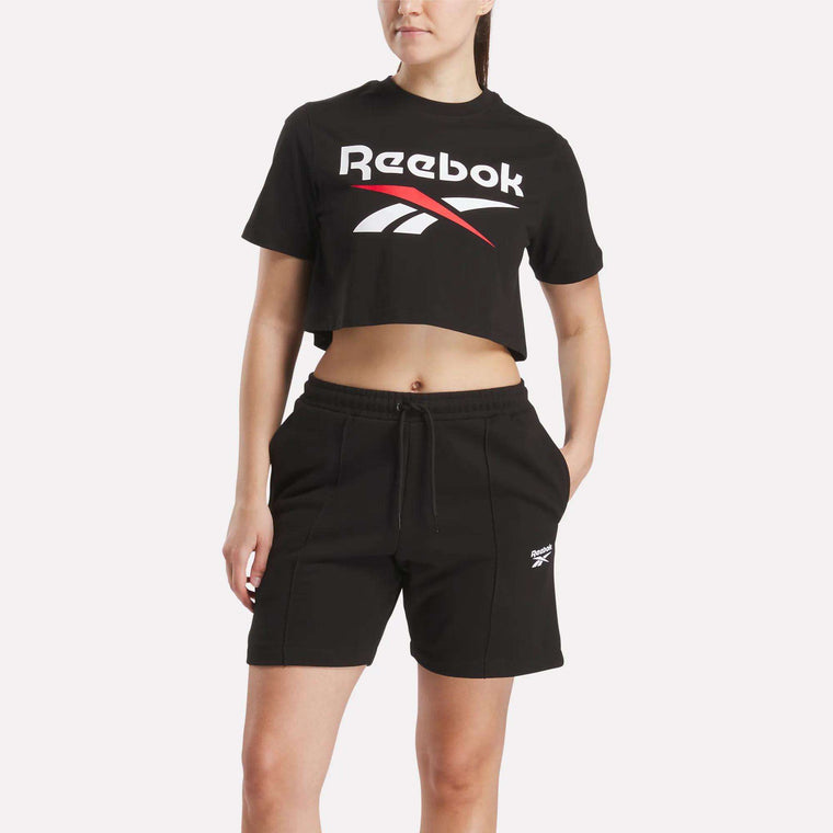 A woman wears the Reebok Women's Two-Color Logo Crew Cropped Short Sleeve Tee and matching shorts, standing with hands in pockets against a plain background.