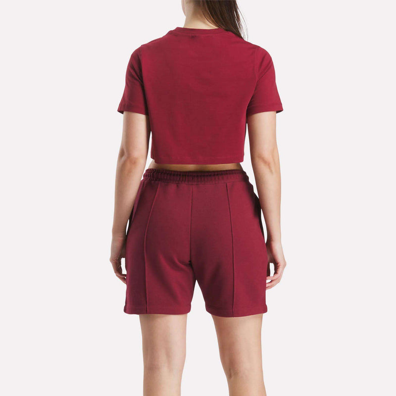 A person faces away from the camera, wearing a maroon Reebok Women's Two-Color Logo Crew Cropped Short Sleeve Tee and matching high-waisted shorts, set against a plain white background.