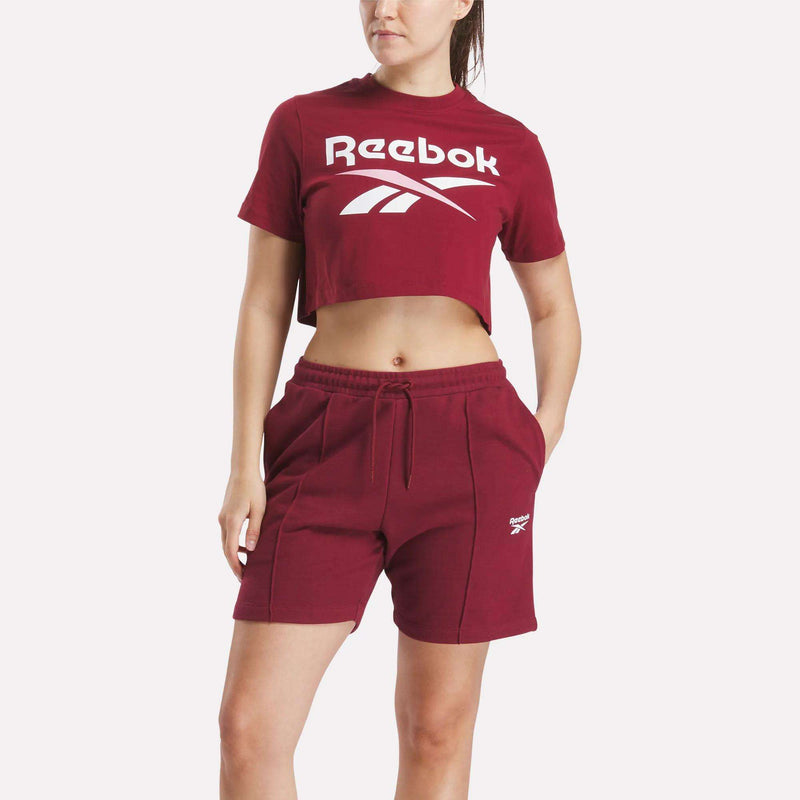 A woman models the Reebok Women's Two-Color Logo Crew Cropped Short Sleeve Tee and matching shorts, both featuring the signature Reebok logo, with one hand in her pocket.