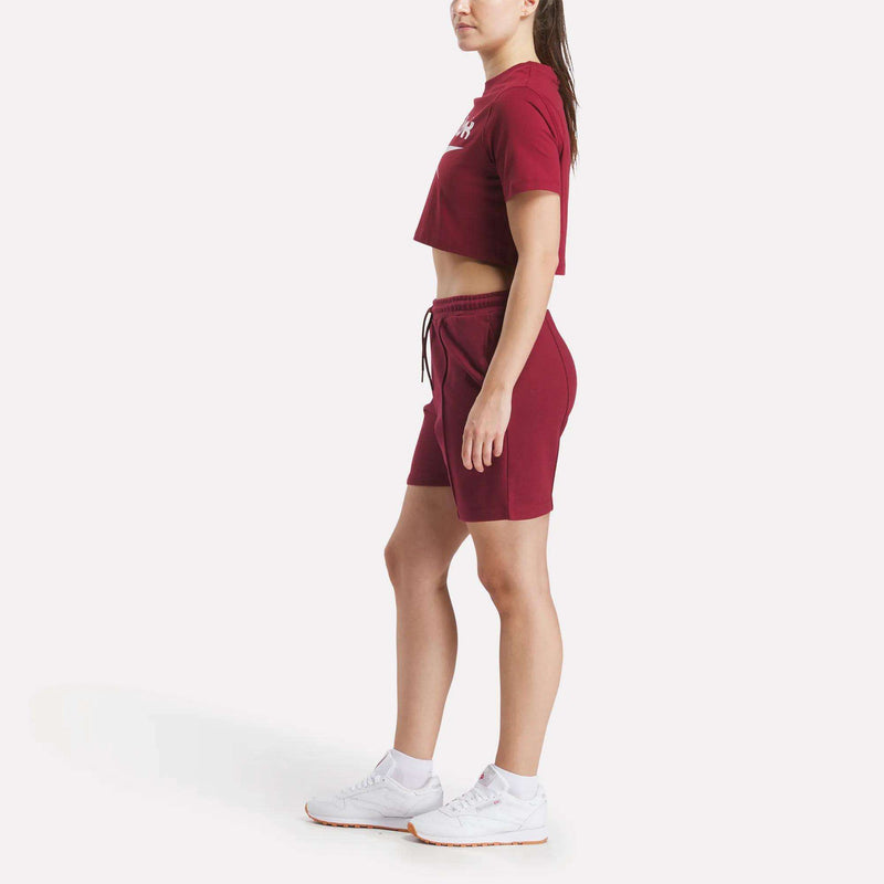 A woman stands sideways in a Reebok Women's Two-Color Logo Crew Cropped Short Sleeve Tee with matching shorts and white sneakers, posed against a plain white background.