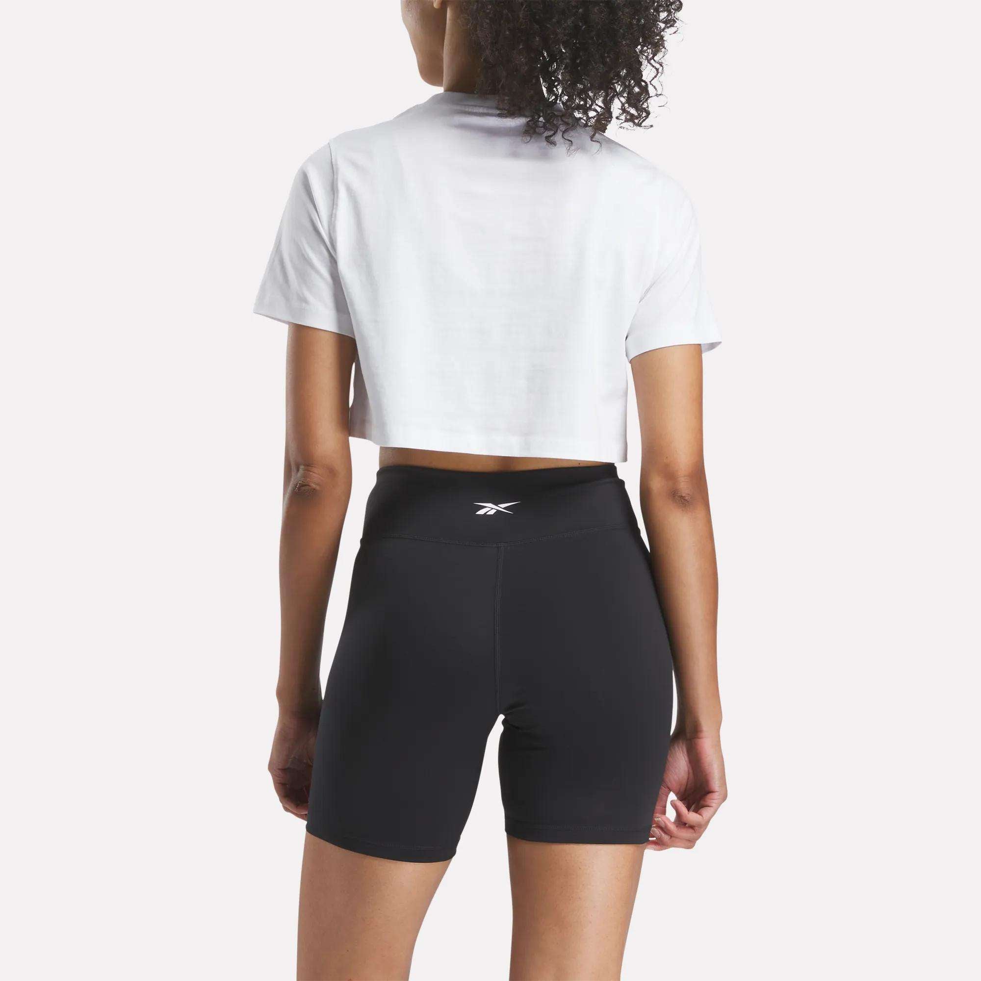 A woman faces away wearing the Reebok Women's Two-Color Logo Crew Cropped Short Sleeve Tee in white, paired with high-waisted black athletic shorts featuring a small logo on the waistband. The background is plain white.