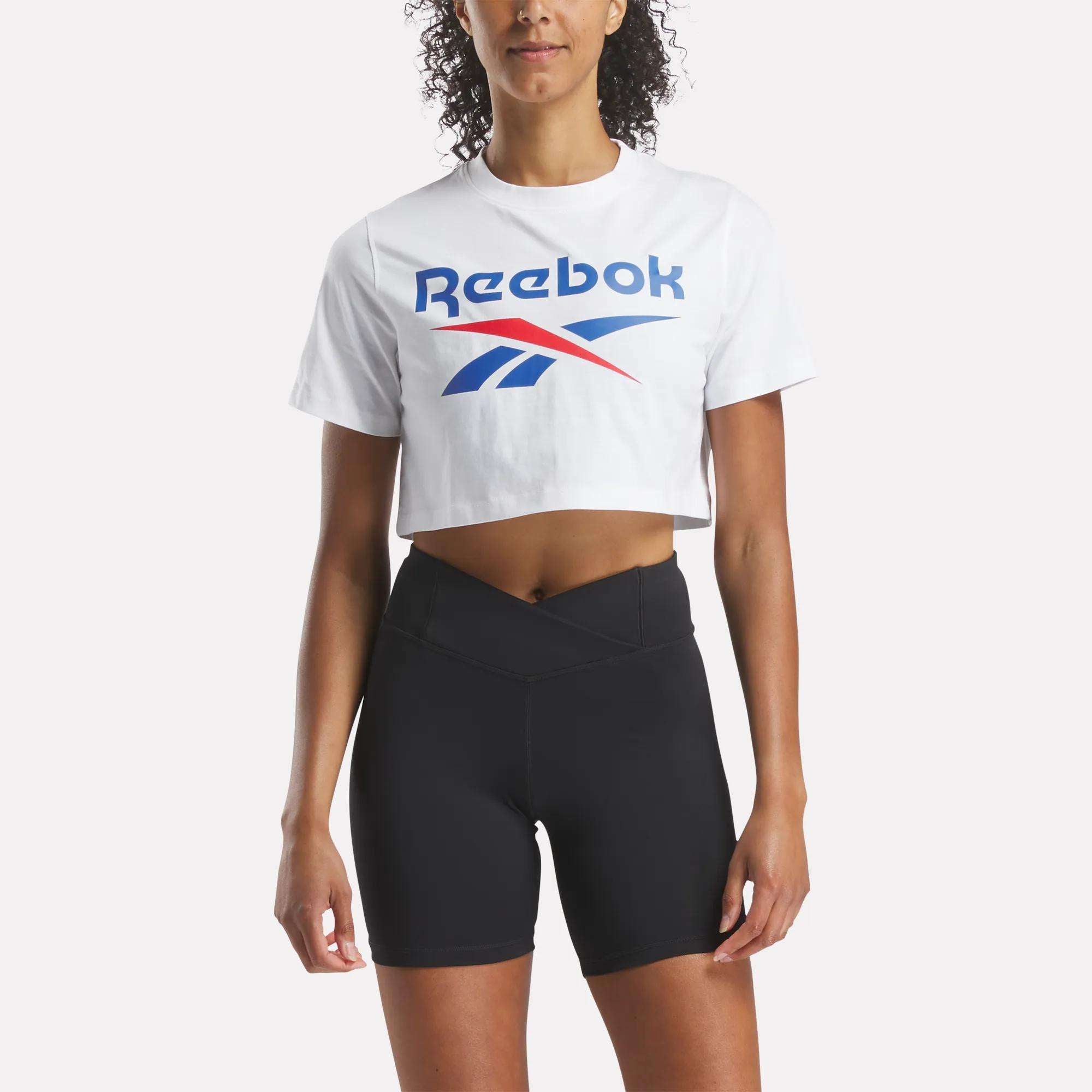 A woman faces forward against a plain light background, wearing a Reebok Women's Two-Color Logo Crew Cropped Short Sleeve Tee in white and high-waisted black cycling shorts.