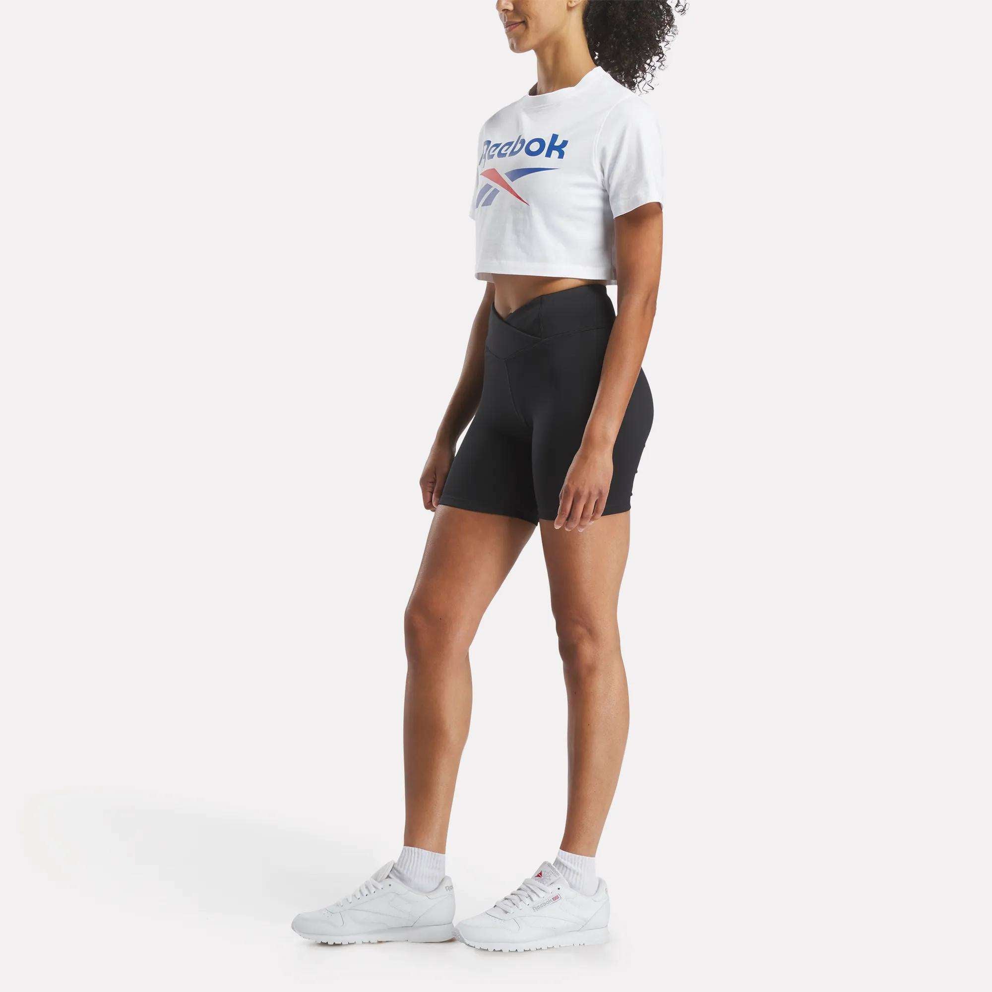 A woman stands sideways against a white background wearing the Reebok Women's Two-Color Logo Crew Cropped Short Sleeve Tee, black high-waisted shorts, white socks, and white sneakers.