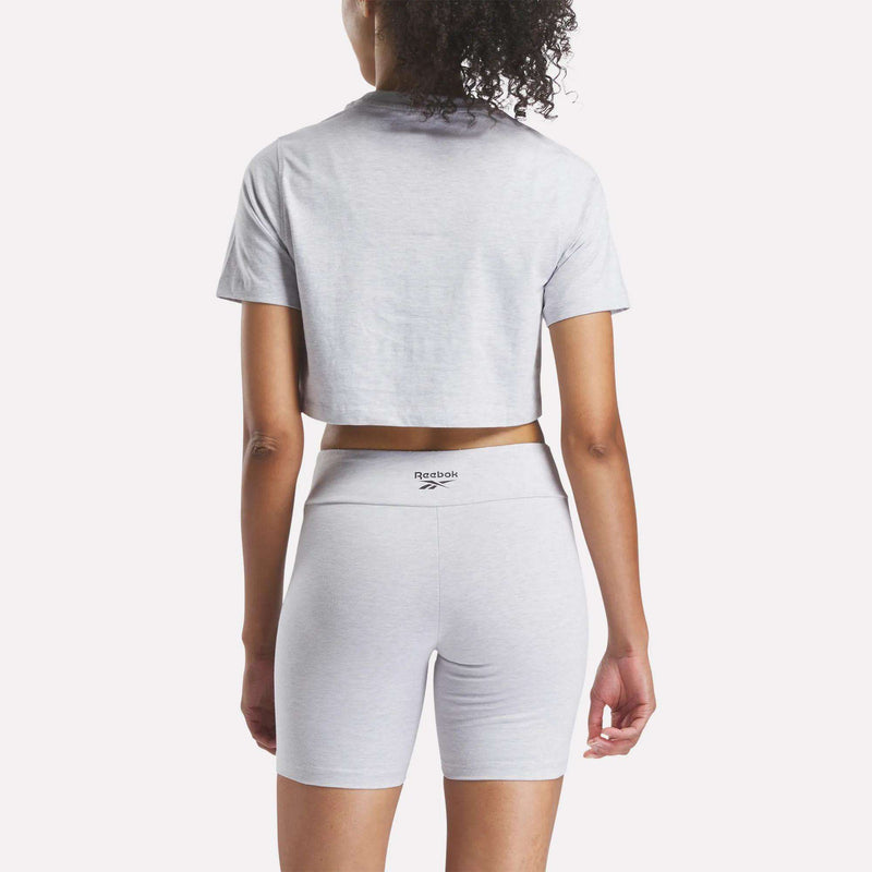 A woman with curly hair is seen from the back, wearing a Reebok Women's Two-Color Logo Crew Cropped Short Sleeve Tee in light gray and matching high-waisted biker shorts with a logo on the waistband.