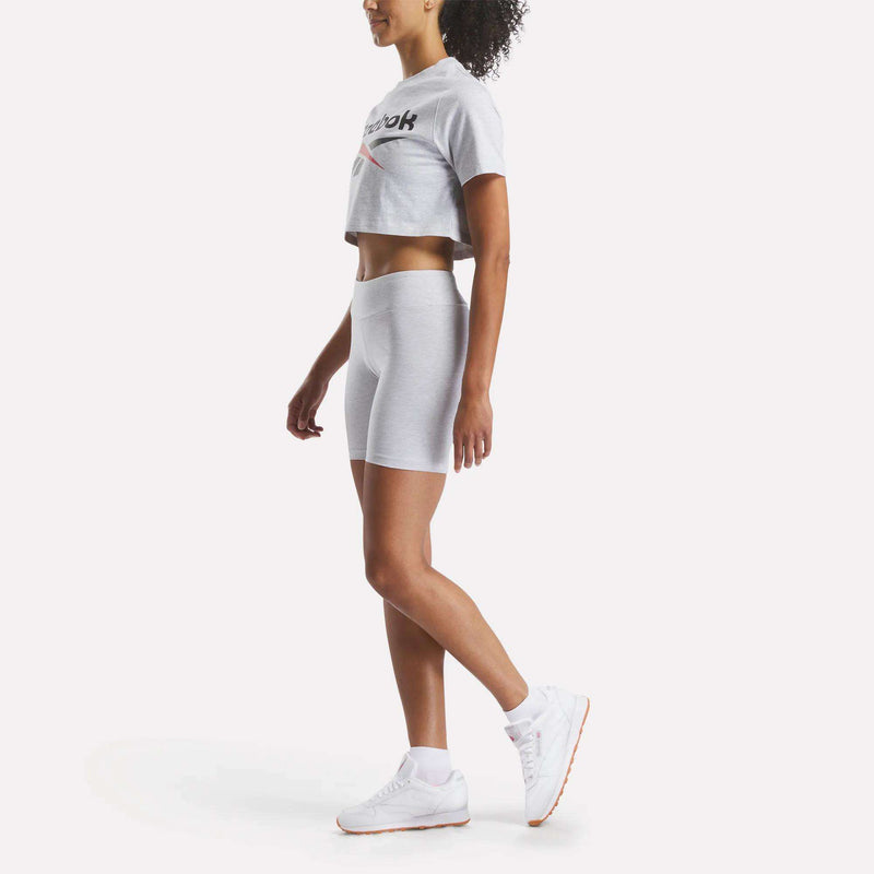 A woman poses sideways in a Reebok Women's Two-Color Logo Crew Cropped Short Sleeve Tee, with matching shorts and white sneakers. Her curly hair is tied back as she stands against a white background.
