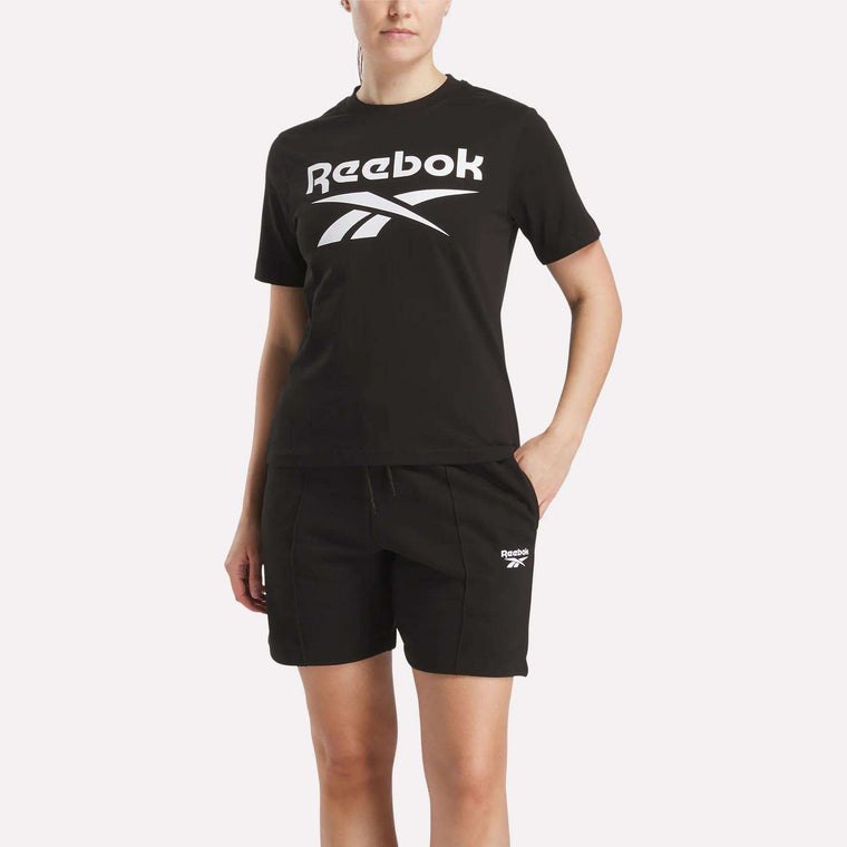A woman in a black Reebok Women’s Big Logo Crewneck Short Sleeve Tee with a bold white Vector logo and matching black shorts stands with one hand in her pocket against a plain light background.