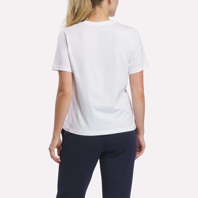 A woman with long blonde hair, dressed in a plain white Reebok Women’s Logo Pocket T-Shirt and dark pants, stands facing away from the camera against a light background.