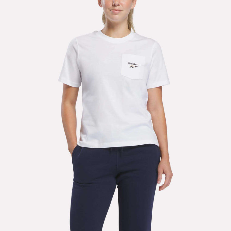A person wearing the Reebok Women’s Logo Pocket T-Shirt in white, featuring a retro Reebok logo and pocket detail, paired with navy pants, stands with one hand in a pocket against a plain white background.