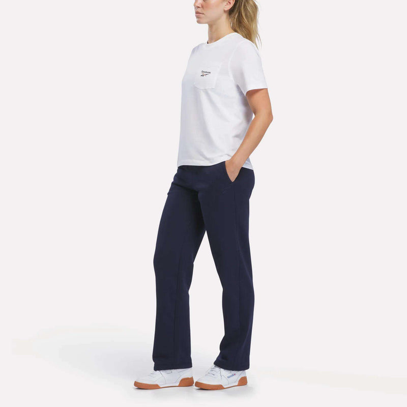 A woman stands in a relaxed pose against a plain white background, wearing the Reebok Women’s Logo Pocket T-Shirt with the retro Reebok logo. She pairs the boxy crop tee with navy blue pants and white sneakers. Her hands rest in her pockets, and her hair is tied back in a ponytail.