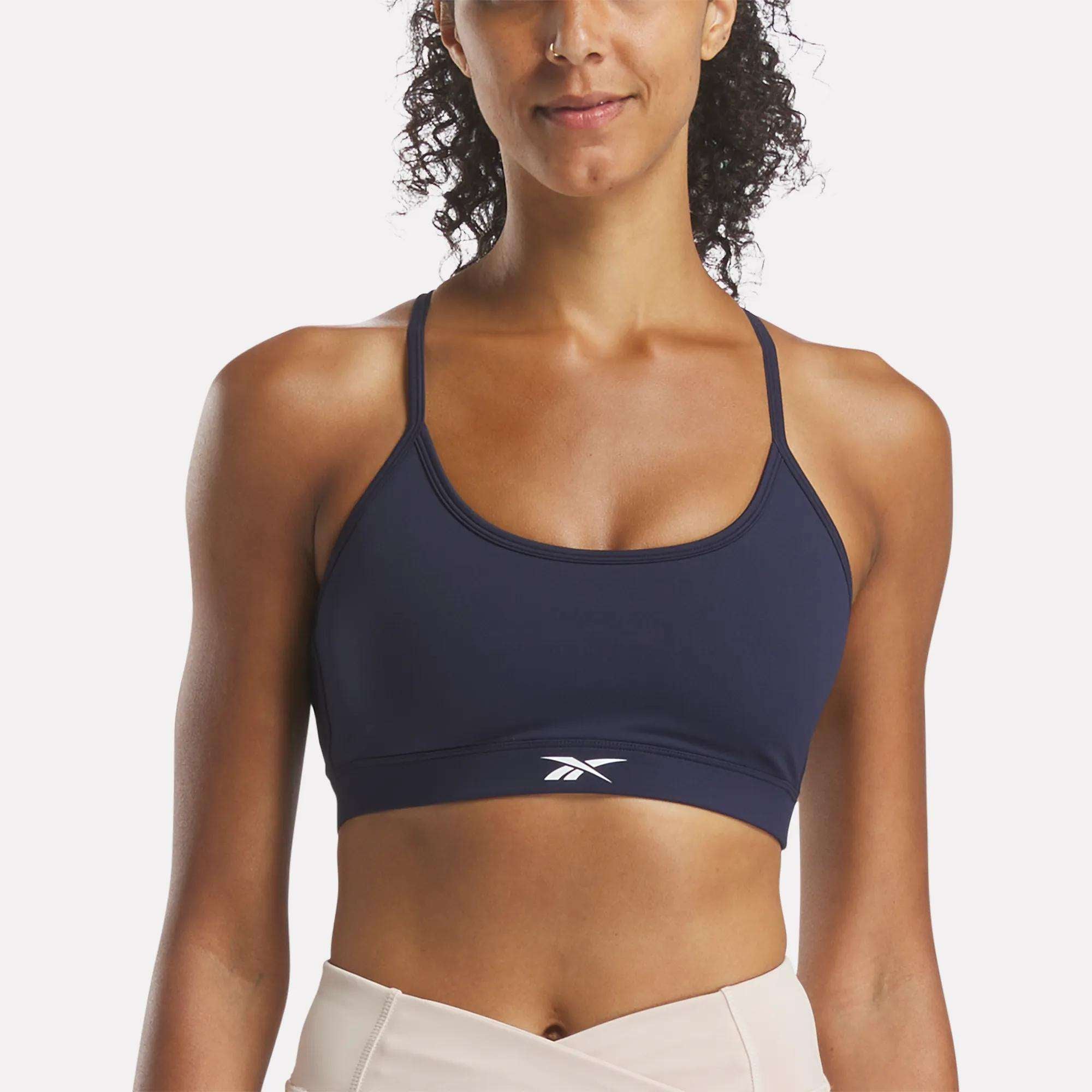 A woman models the Reebok Women's Core Strappy Bra - Medium Support in navy blue, featuring thin straps and a white logo on the front, ideal for medium-impact workouts, against a plain background.