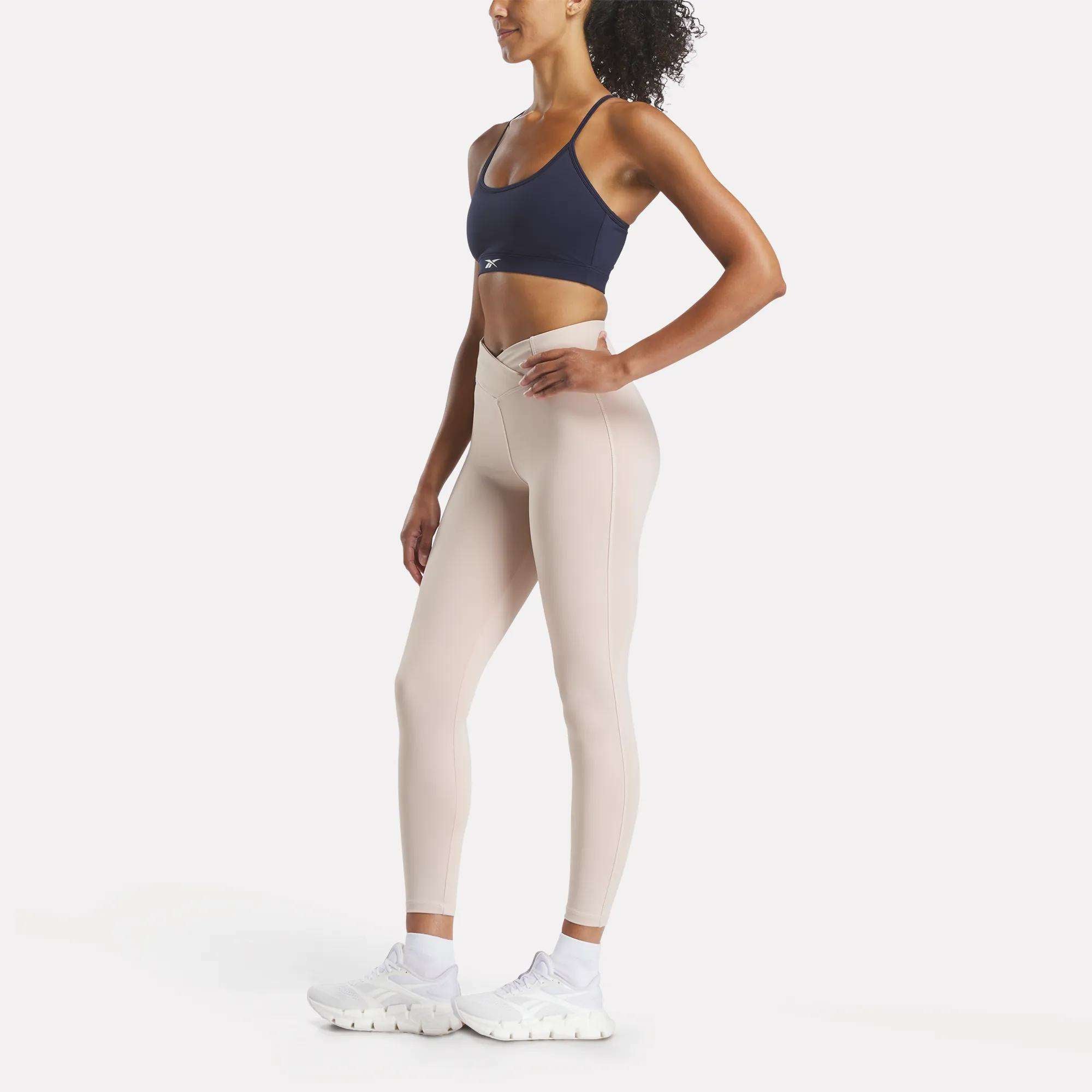 A woman poses sideways in a Reebok Women's Core Strappy Bra - Medium Support (navy), light beige leggings, and white athletic shoes, set against a plain white background.