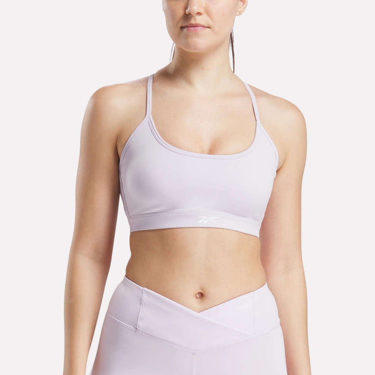 A woman wears the Reebok Women's Core Strappy Bra - Medium Support in light purple, paired with matching high-waisted leggings. She stands against a white background. The outfit is ideal for medium-impact workouts and features quick-drying fabric.