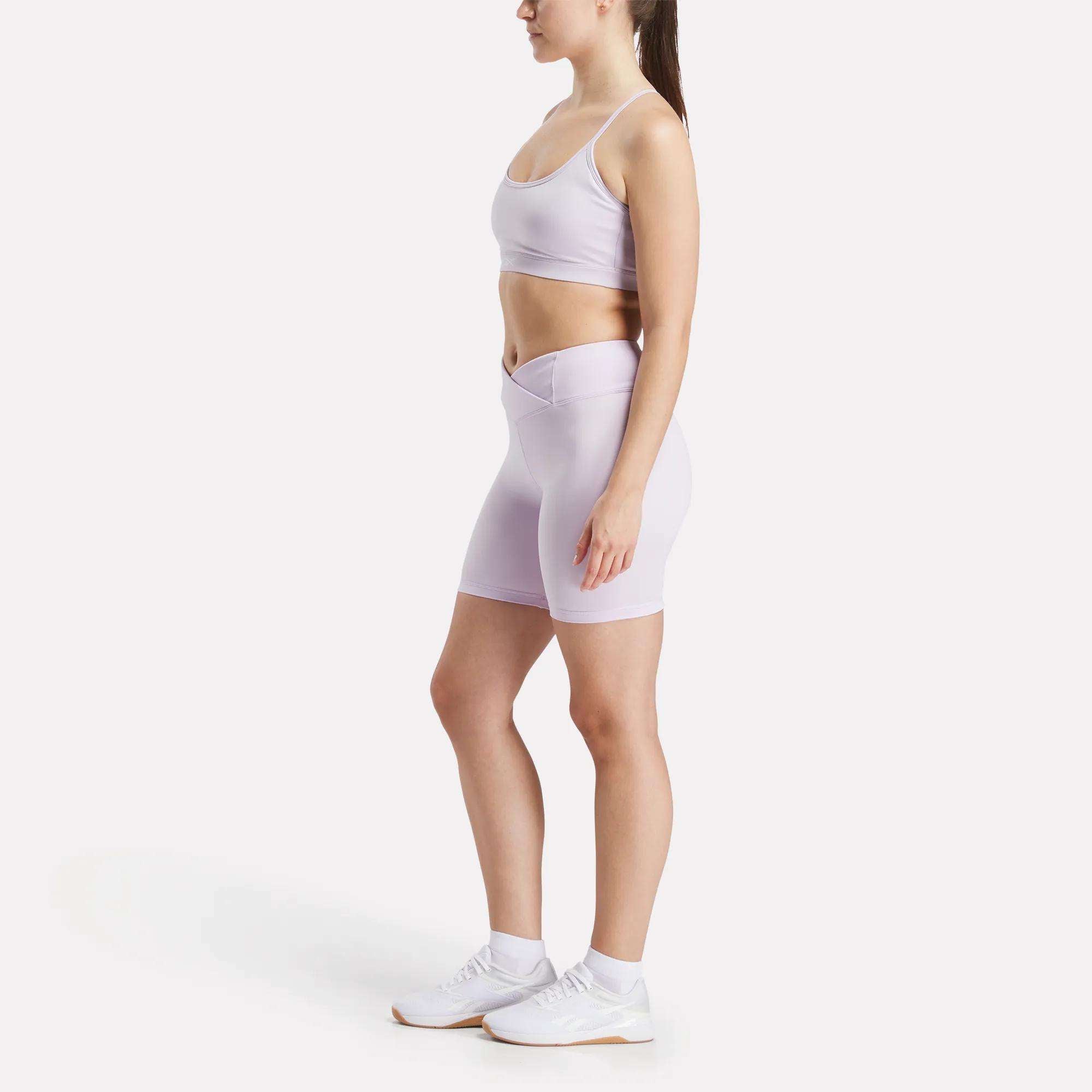 A woman stands in profile wearing the Reebok Women's Core Strappy Bra - Medium Support in light purple, paired with matching high-waisted bike shorts, white socks, and sneakers, against a plain white background.