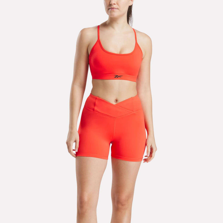 A woman wears a Reebok Women's Strappy Sports Bra in bright orange with matching high-waisted shorts. The sports bra, ideal for medium-impact workouts, features quick-drying fabric and displays the Reebok logo on the front.