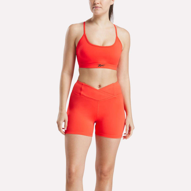 A woman wears a Reebok Women's Strappy Sports Bra in bright orange with matching high-waisted shorts. The sports bra, ideal for medium-impact workouts, features quick-drying fabric and displays the Reebok logo on the front.