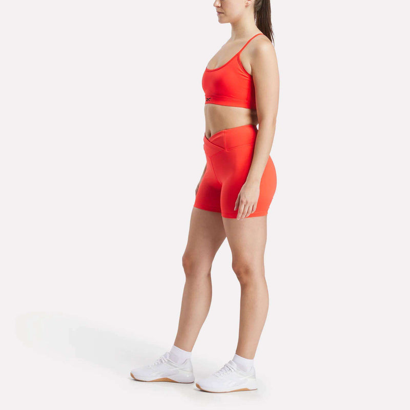 A woman stands in profile against a plain white background wearing the Reebok Women’s Strappy Sports Bra and matching bright orange shorts set, paired with white socks and sneakers—an ideal choice for medium-impact workouts thanks to its quick-drying fabric.