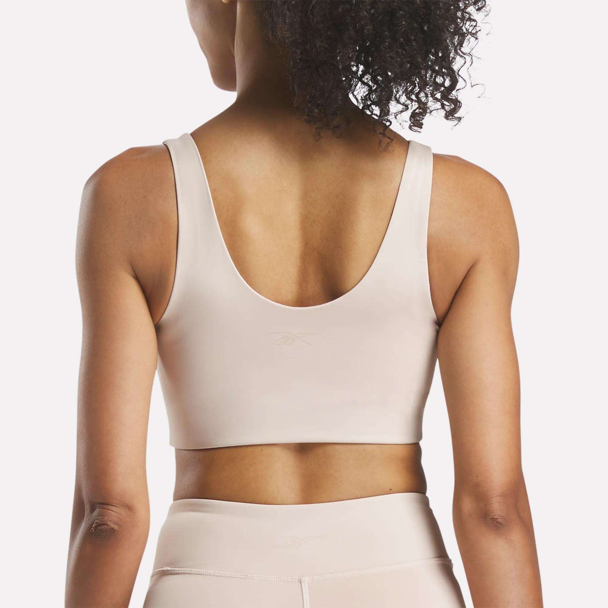 A woman with curly hair is seen from behind wearing a light beige Reebok Women's Yoga V-Neck Bra and matching high-waisted leggings, ideal for studio workouts, against a plain background.