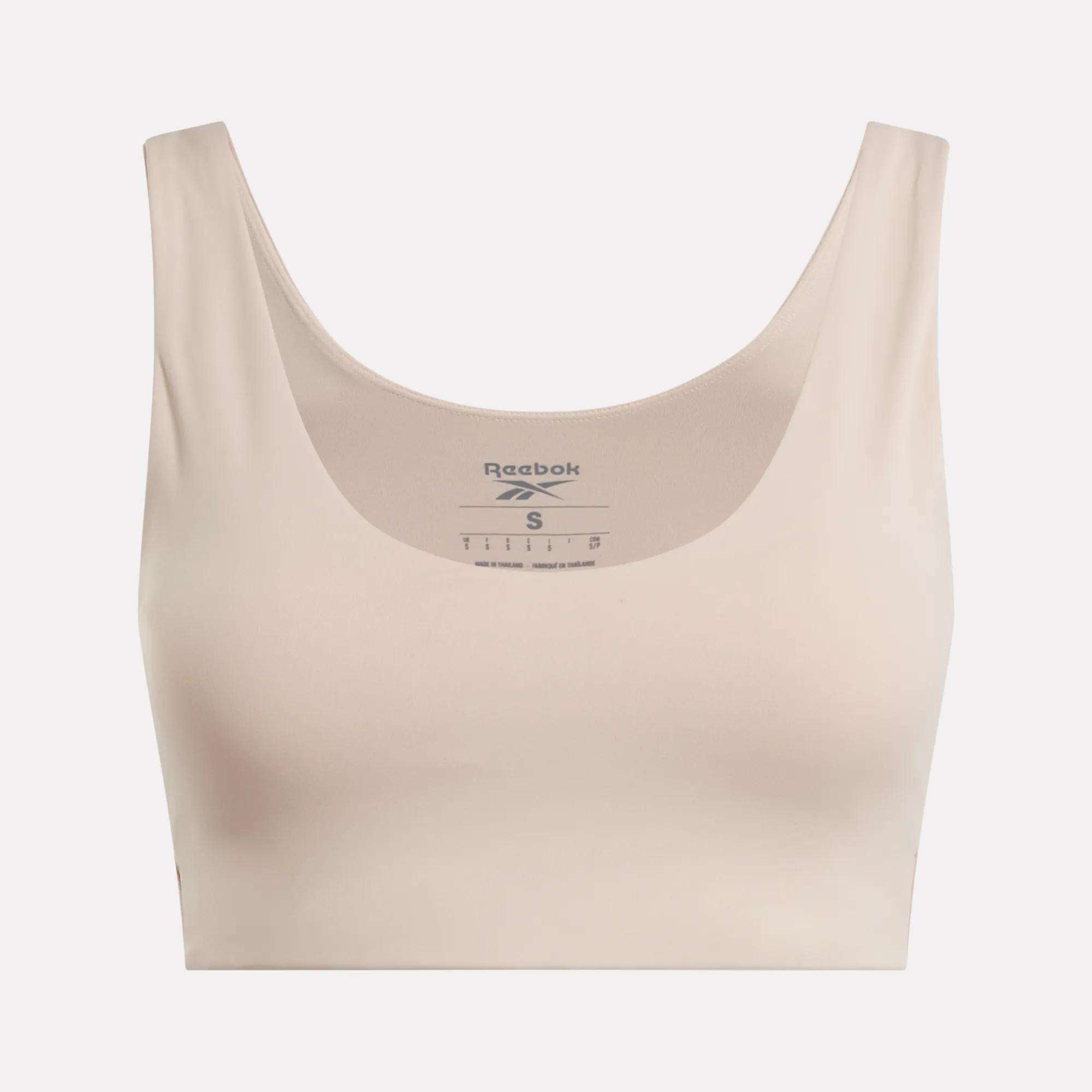 The Reebok Women's Yoga V-Neck Bra in beige features a scoop neckline and wide straps, offering medium-impact support for studio workouts. The Reebok logo and sizing appear inside the neckline on a plain light background.