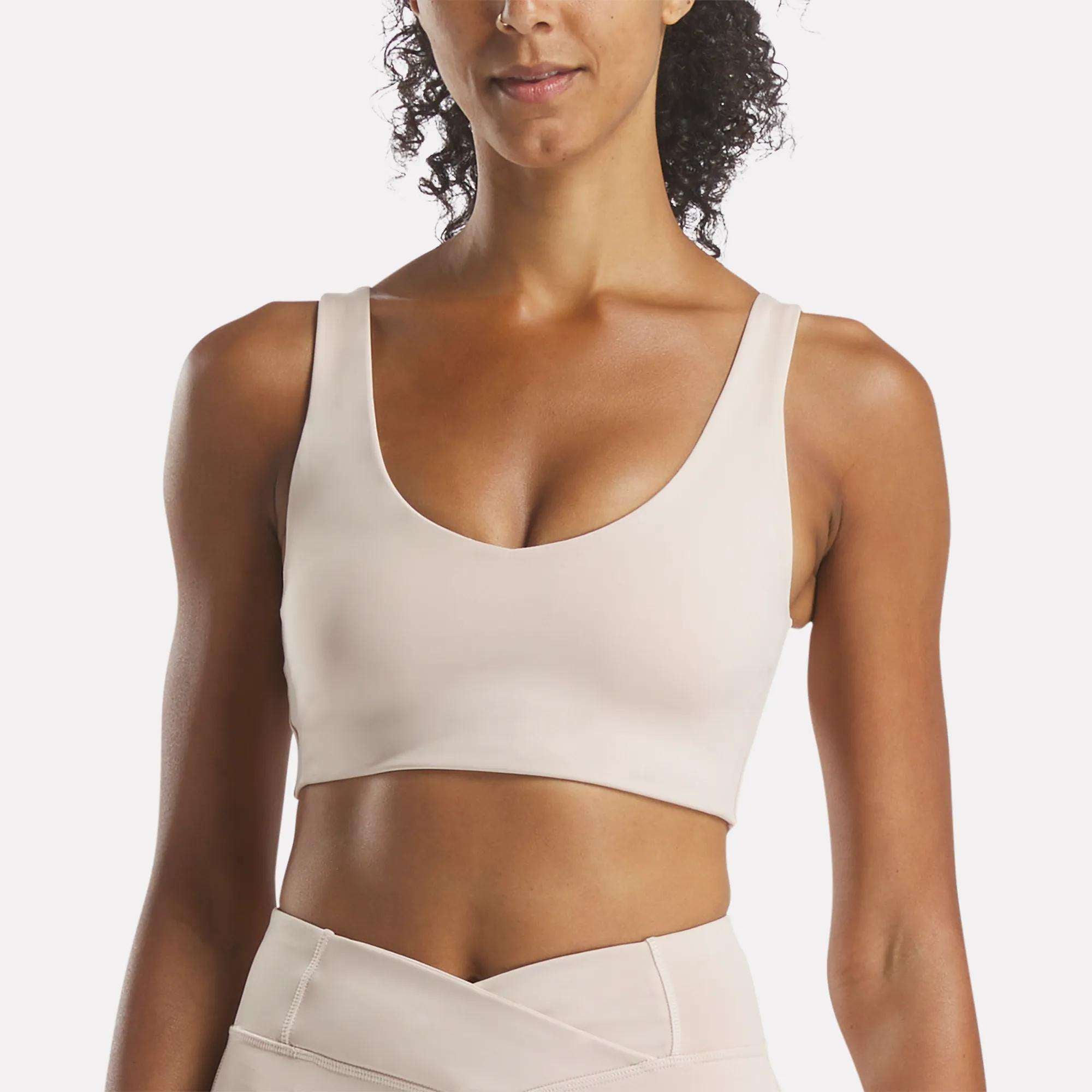 A woman models the Reebok Women's Yoga V-Neck Bra in light beige with medium-impact support and matching high-waisted leggings, shown from shoulders to waist against a white background—ideal for studio workouts.
