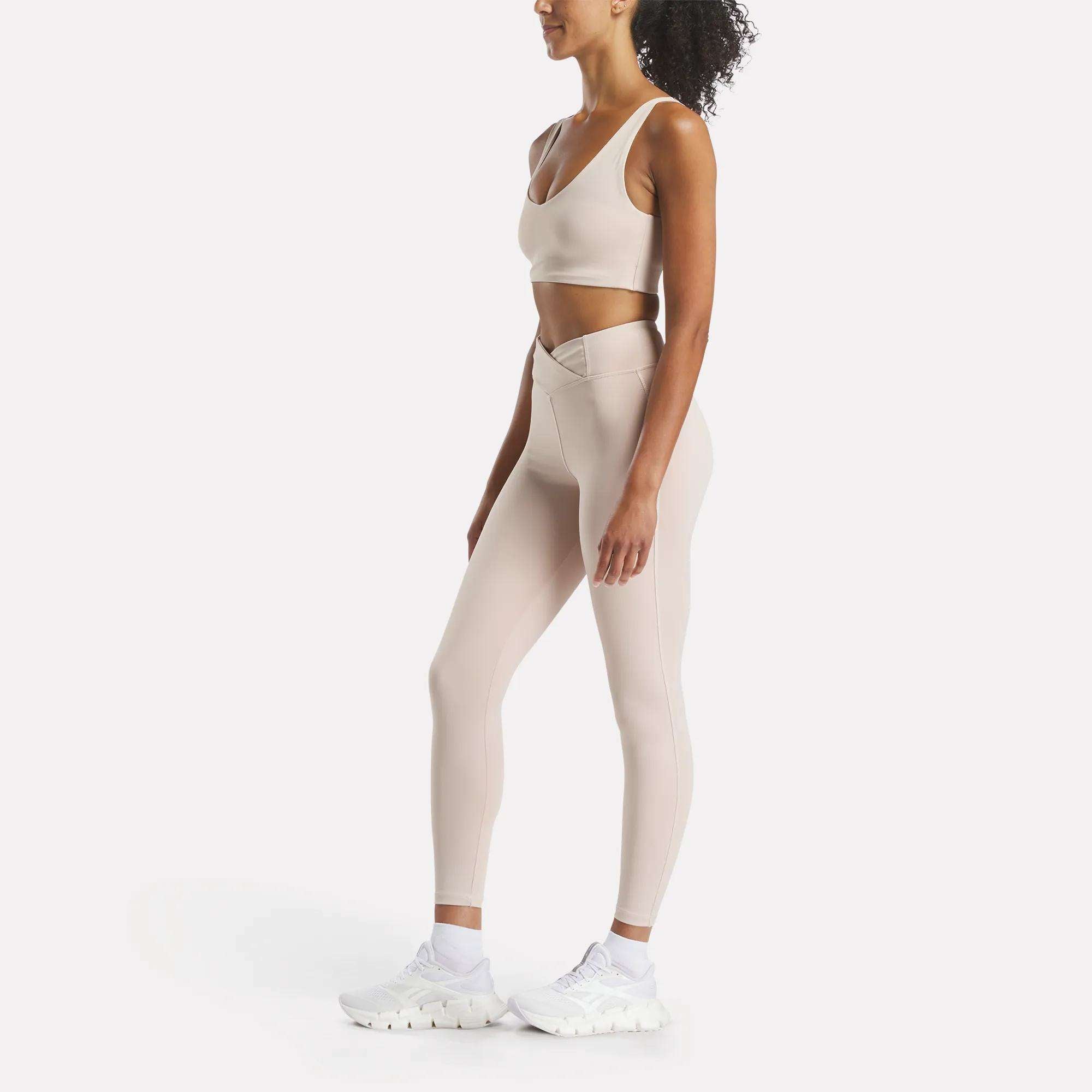 Woman in profile wears a light beige Reebok Women's Yoga V-Neck Bra and matching high-waisted leggings with a V-shaped waistband, paired with white athletic shoes—an ideal look for studio workouts—posed against a plain white background.