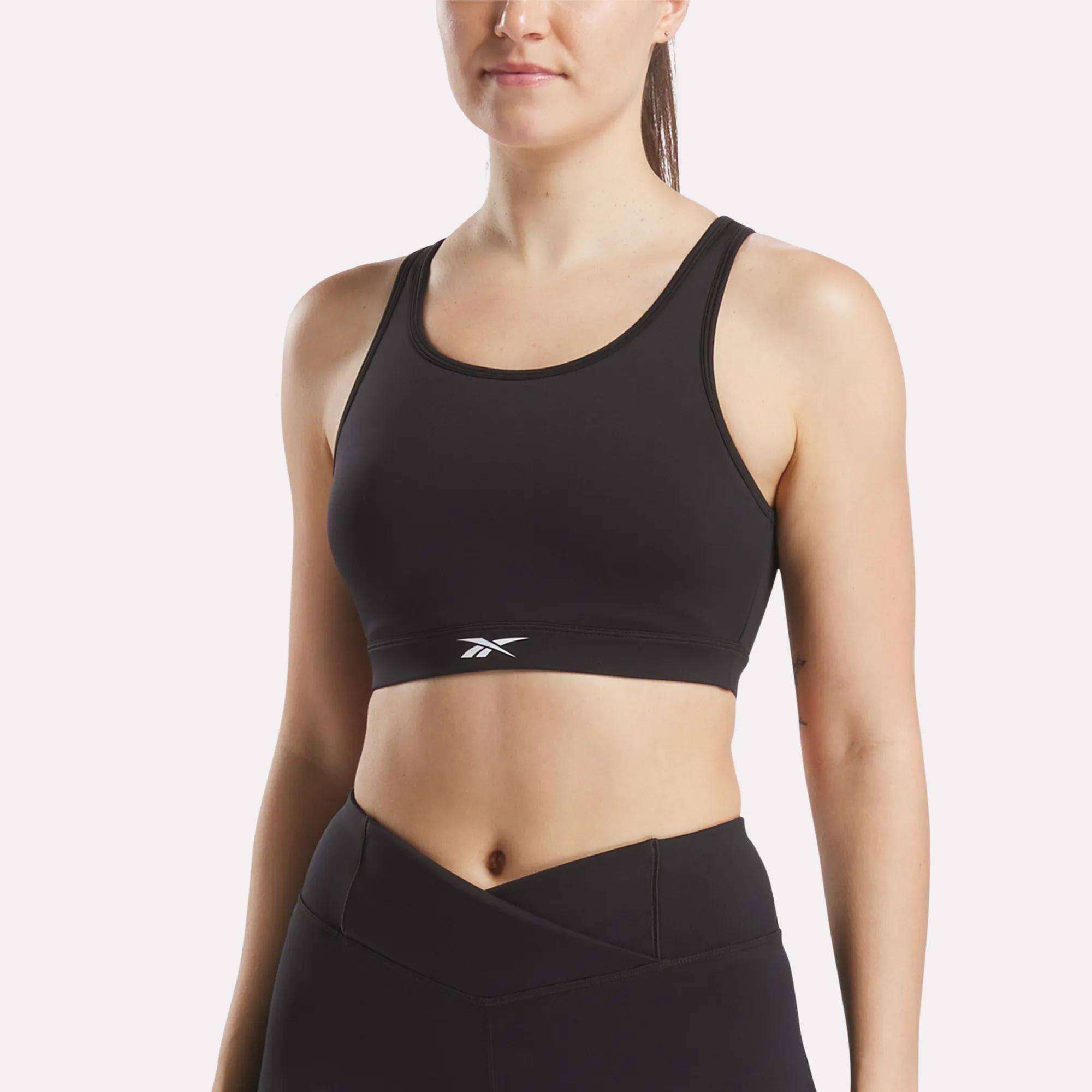 A woman in a black Reebok Women's Core High-Impact Bra with a small white logo and matching high-waisted leggings stands against a plain white background. The sweat-wicking bra offers support and style.