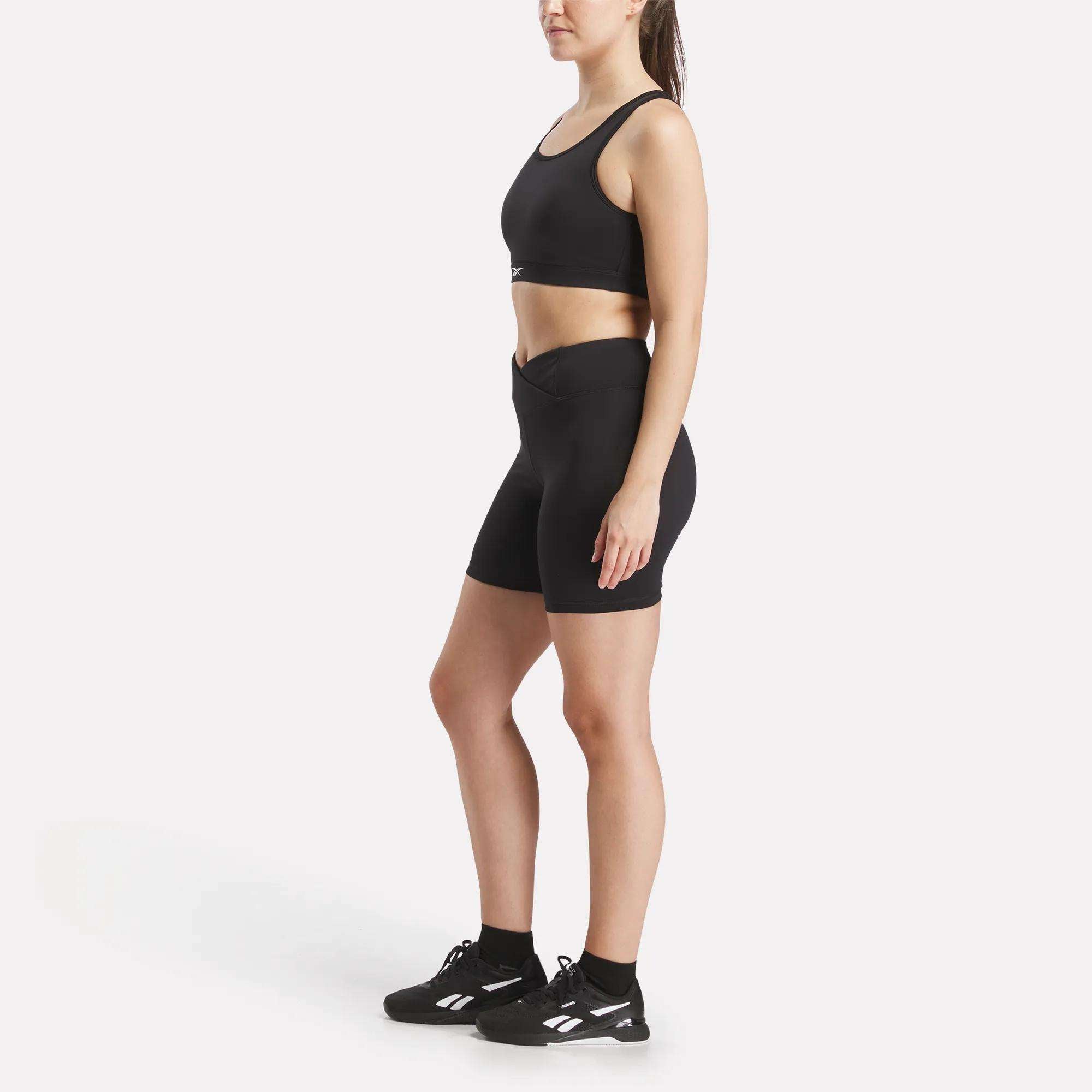 A woman stands sideways against a plain white background wearing the Reebok Women's Core High-Impact Bra, high-waisted black athletic shorts, and black sneakers with white accents.