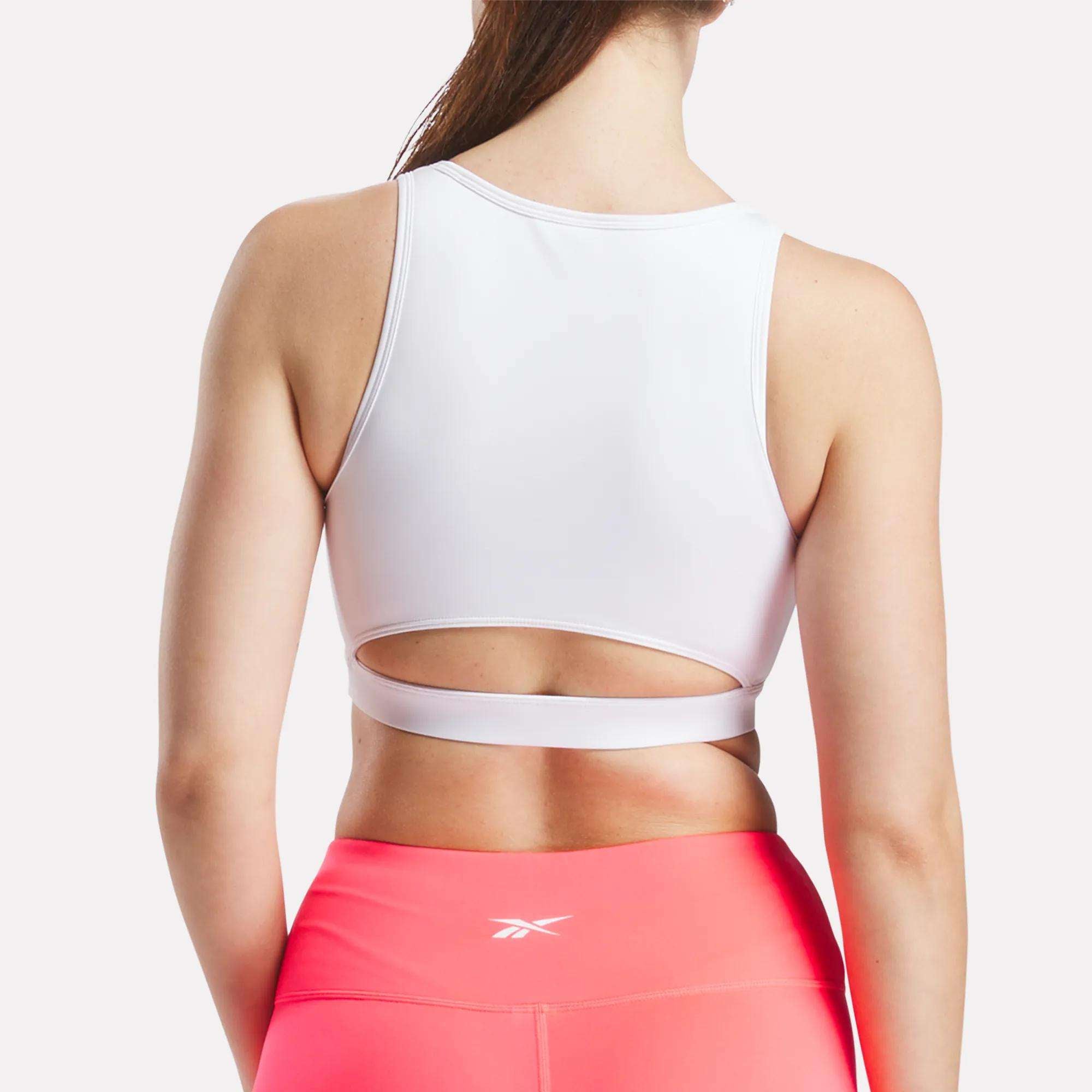 A woman stands with her back to the camera, wearing Reebok's Women's Core High-Impact Bra in white, featuring a cutout design, and bright pink leggings. Her long hair flows down against a plain light background.