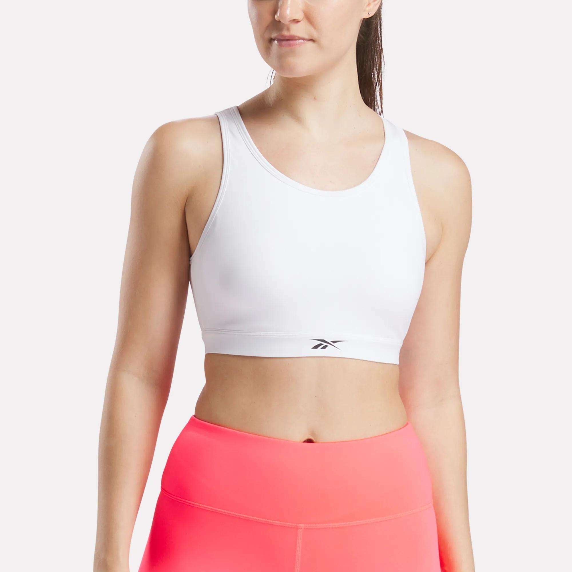 A woman wears the Reebok Women's Core High-Impact Bra and high-waisted neon pink workout leggings, standing against a plain light background with her face partially out of frame.