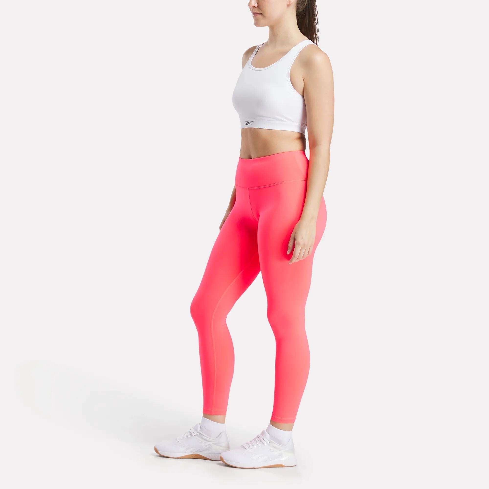 A woman models the Reebok Women's Core High-Impact Bra with bright pink leggings and white sneakers, standing relaxed in a side-facing pose against a plain white background.