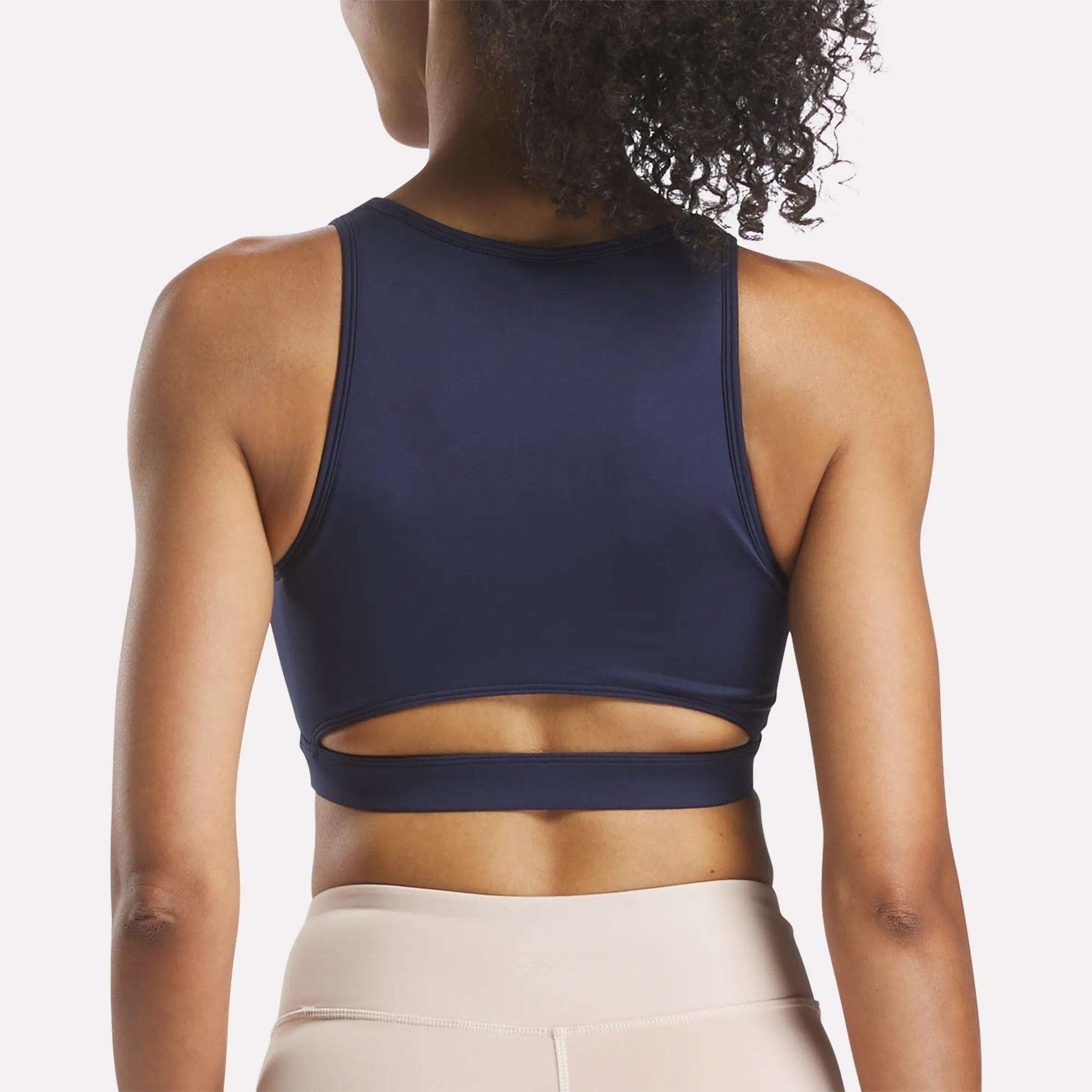 A woman with curly hair models the Reebok Women's Core High-Impact Bra in navy blue, featuring a lower back cutout, paired with light beige leggings. Shown from behind against a white background. Ideal for high-impact training.