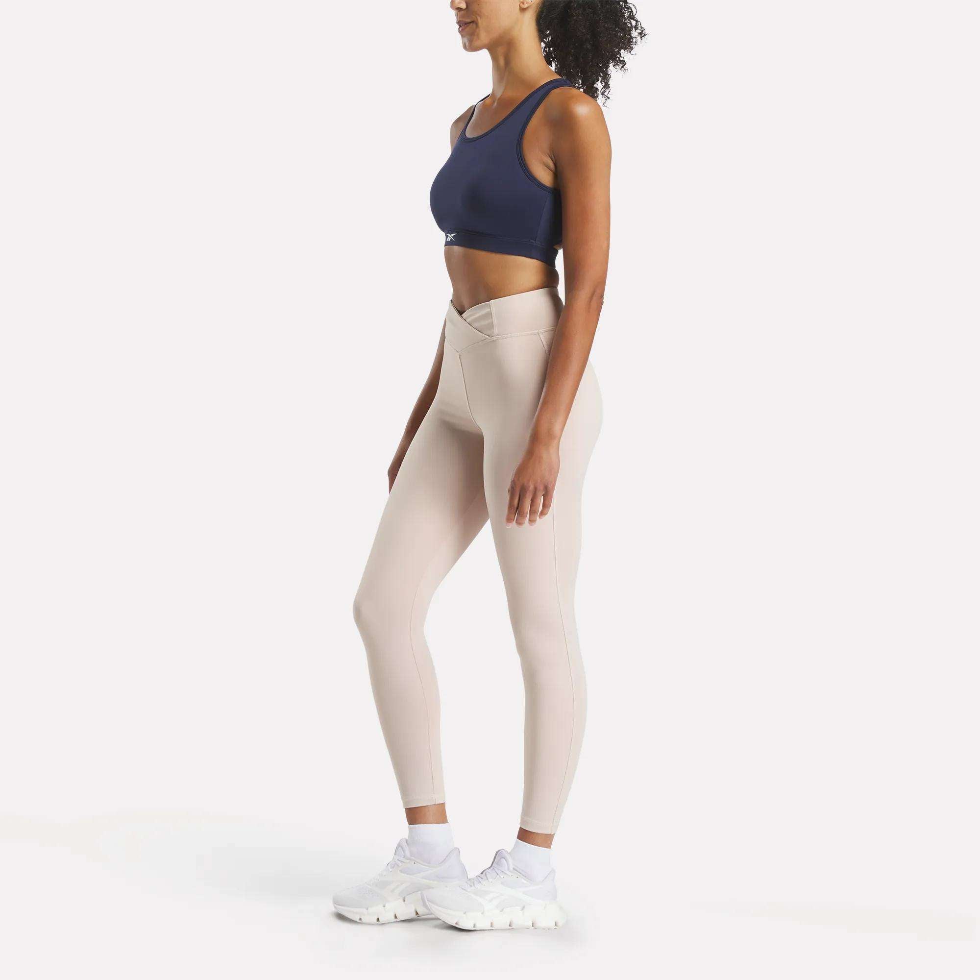 A woman stands in profile wearing a Reebok Women's Core High-Impact Bra in navy, high-waisted beige leggings with a V-shaped waistband, and white sneakers against a plain white background.