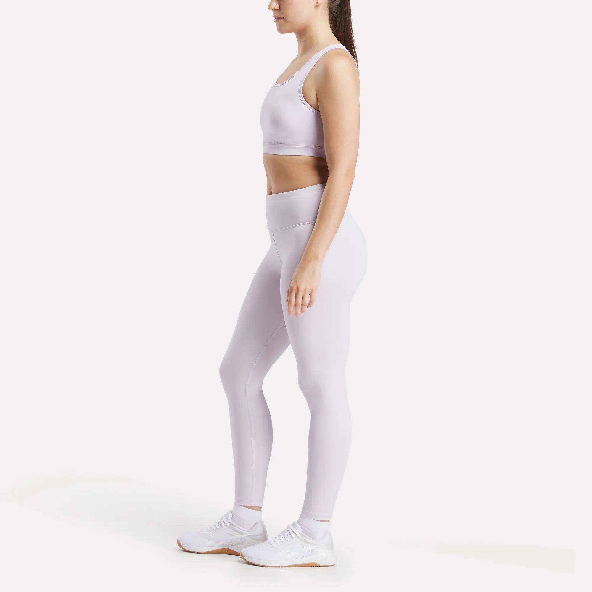 A woman stands in profile against a white background, wearing the Reebok Women's Core High-Impact Bra and matching high-waisted leggings in light purple with white sneakers—ideal gear for high-impact training.