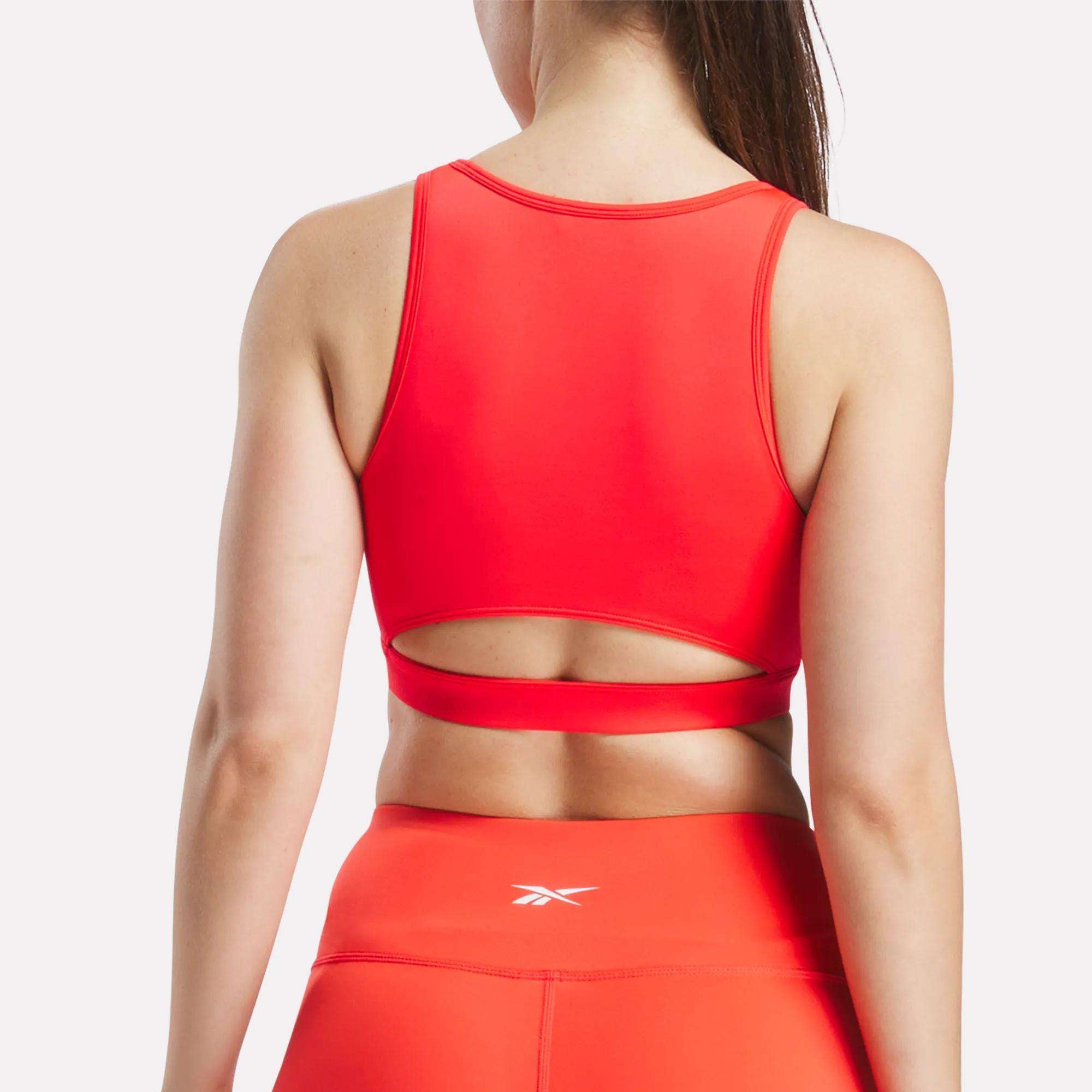 A woman models the Reebok Women's Core High-Impact Bra in bright red with a back cutout, paired with matching high-waisted leggings, seen from behind against a white background.