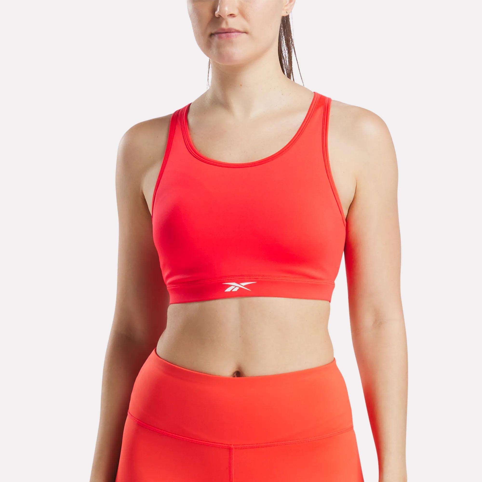 A woman wears the Reebok Women's Core High-Impact Bra, a bright orange racerback sports bra with a small white logo on the front, paired with matching high-waisted leggings against a plain, light background.