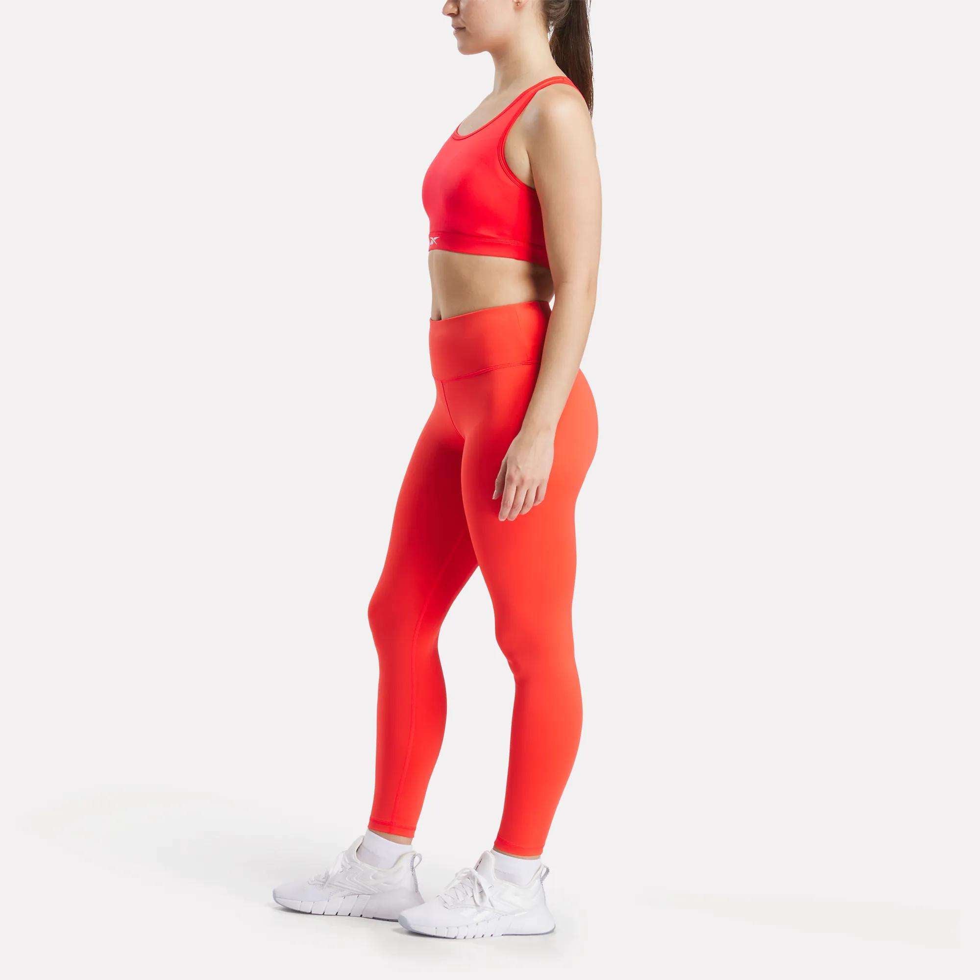 A woman stands in profile against a plain white background, wearing a matching bright red Reebok Women's Core High-Impact Bra and high-waisted leggings, paired with white athletic shoes.