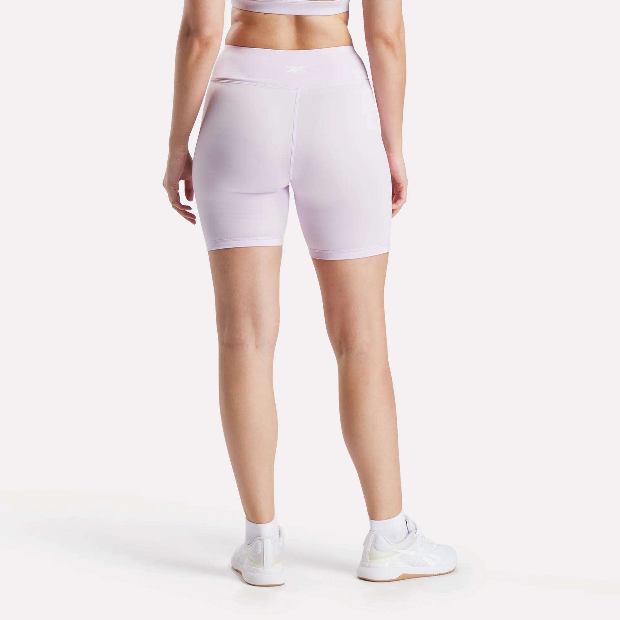 A person stands with their back to the camera against a white background, wearing Reebok Women's Core High-Rise Cycle Shorts in light purple with a matching sports bra, both made from sweat-wicking fabric.