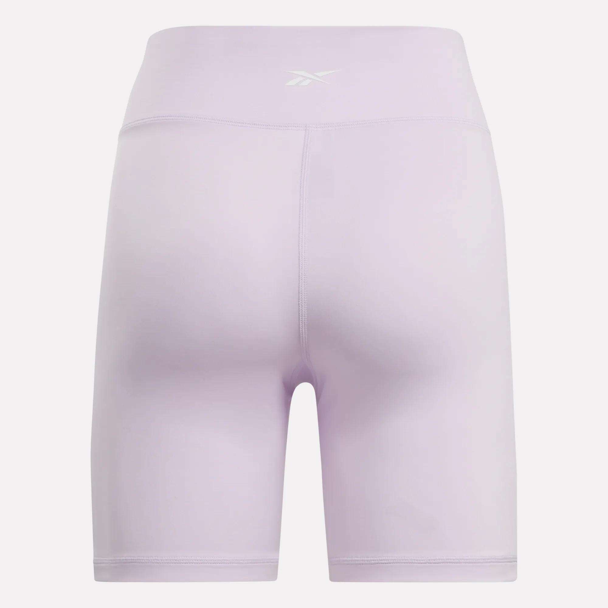 The Reebok Women's Core High-Rise Cycle Shorts in light purple are shown from the back on a white background, featuring a small white Reebok logo on the waistband and sweat-wicking fabric for all-day comfort.