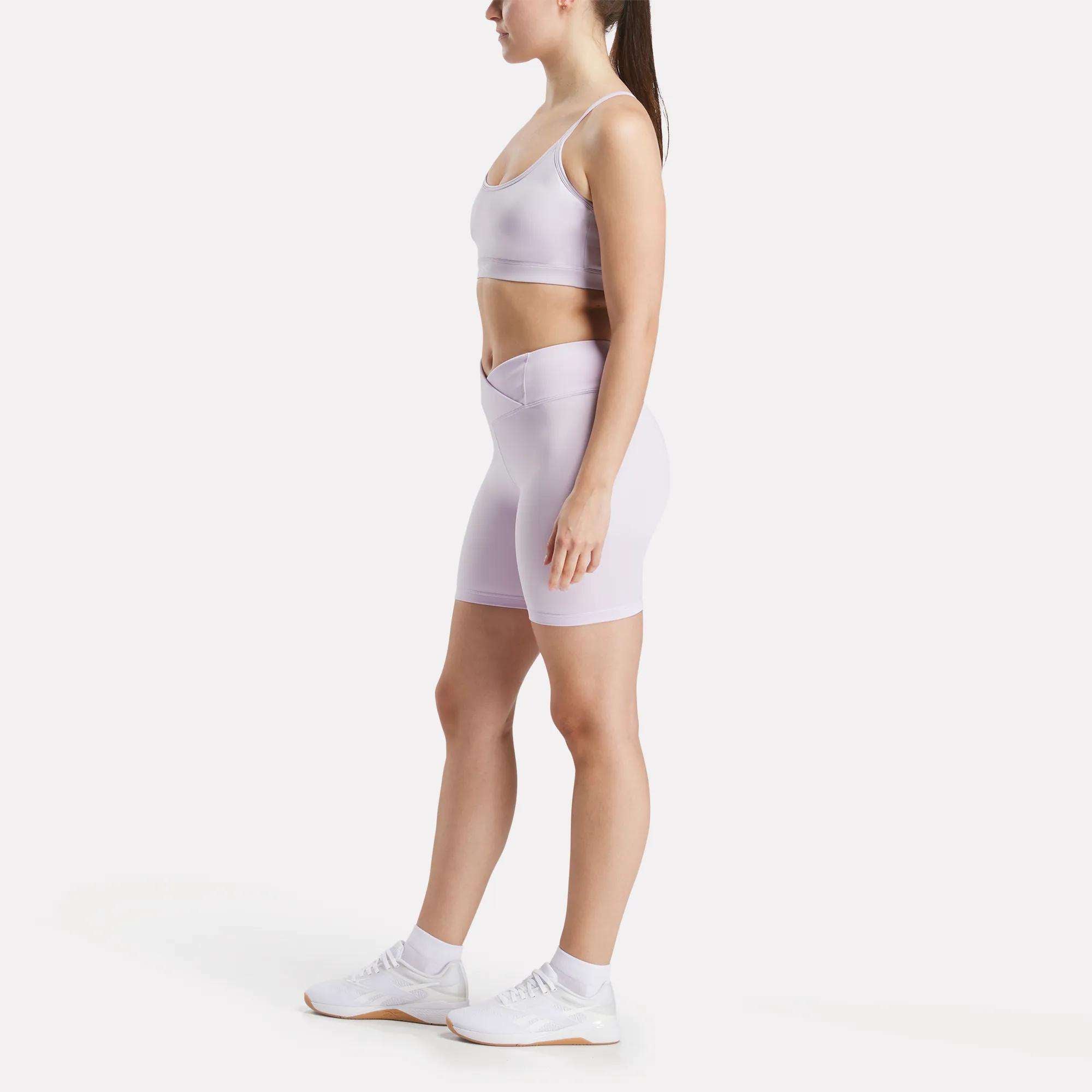 A woman in profile models Reebok Women's Core High-Rise Cycle Shorts and a light purple sports bra with white sneakers, set against a plain white background.