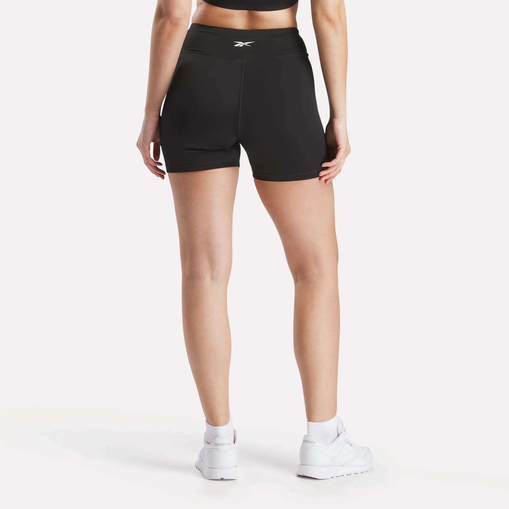 A woman faces away, wearing black Reebok Women's Core High-Rise Shorts with a matching sports bra and white sneakers. These sweat-wicking shorts from Reebok ensure comfort during workouts.