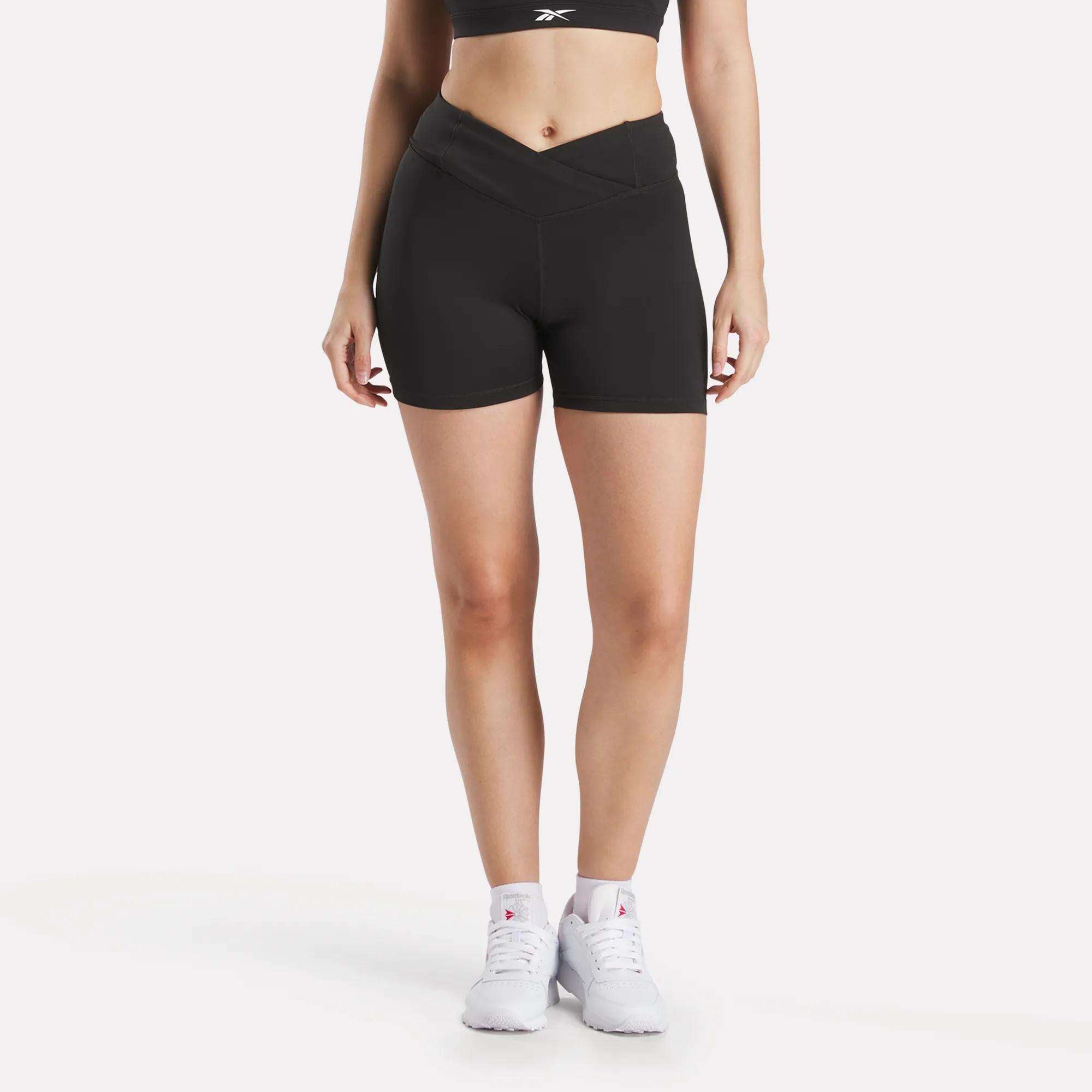 A woman models Reebok Women's Core High-Rise Shorts with a V-shaped waistband and a black sports bra, paired with white sneakers against a plain white background. Her face is not visible.