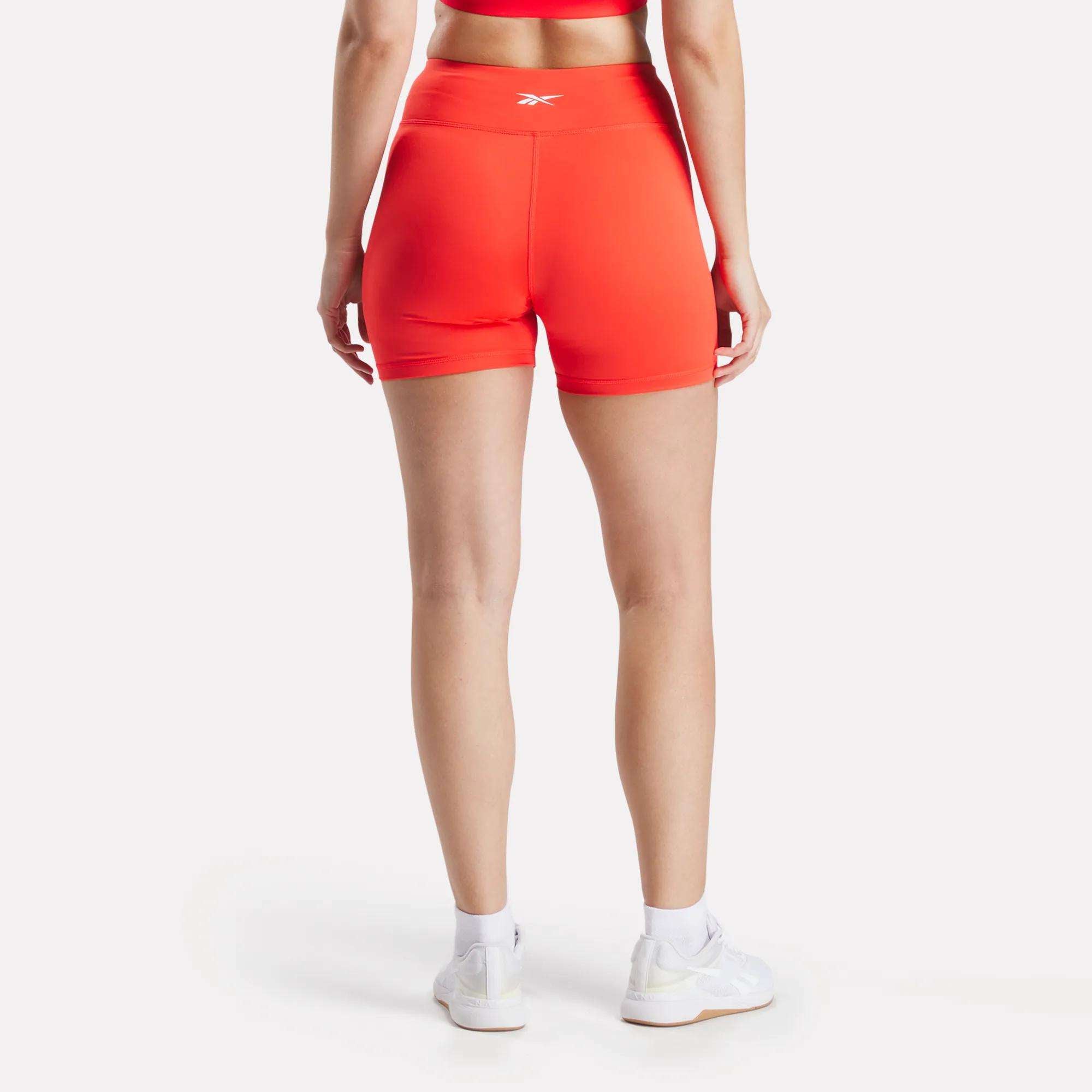 A person faces away, modeling Reebok Women's Core High-Rise Shorts in bright orange with a matching top, paired with white socks and sneakers, against a plain white background.
