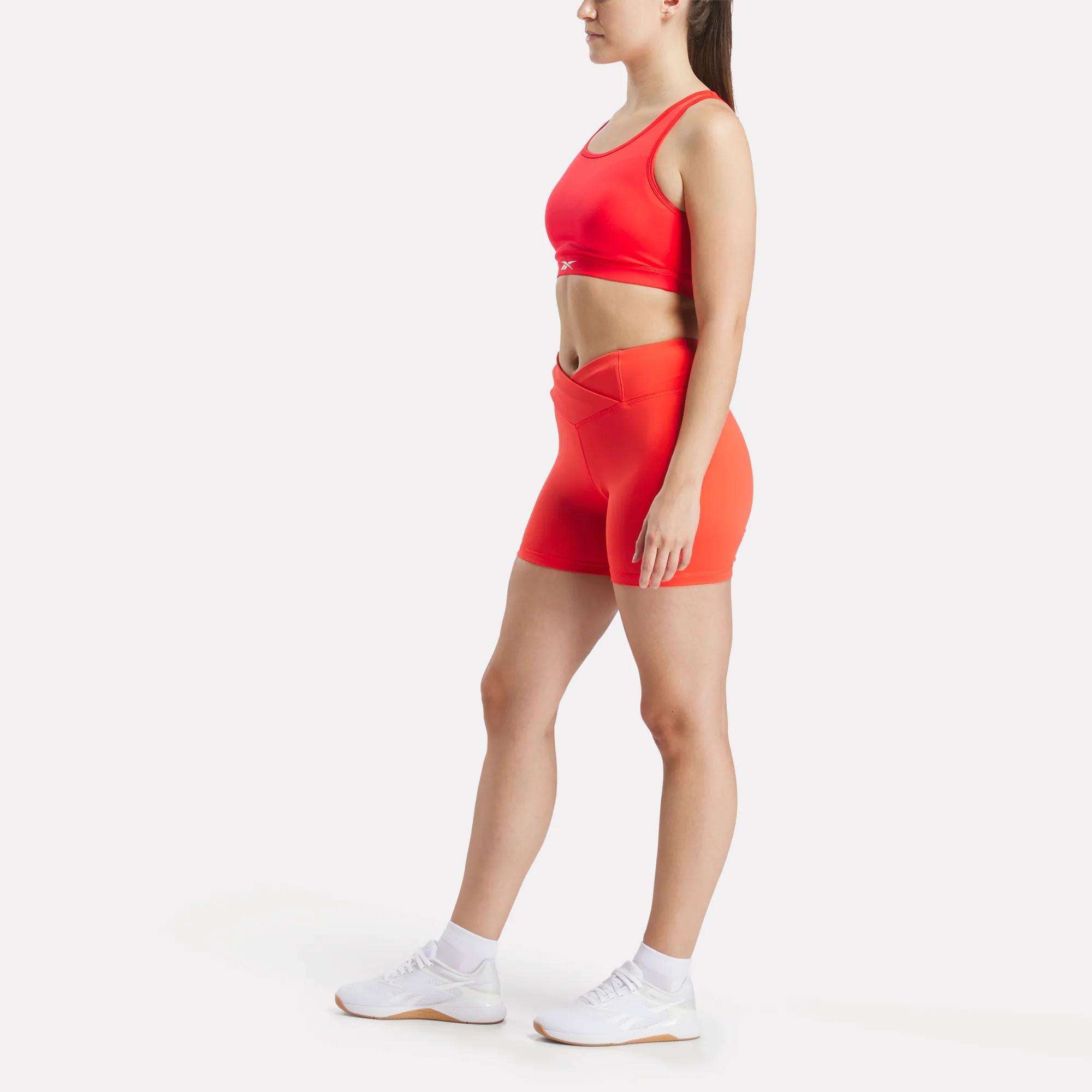 A woman stands against a white background wearing Reebok Women's Core High-Rise Shorts in bright red, paired with a matching sports bra, white ankle socks, and athletic shoes. She faces slightly right with arms relaxed by her sides.