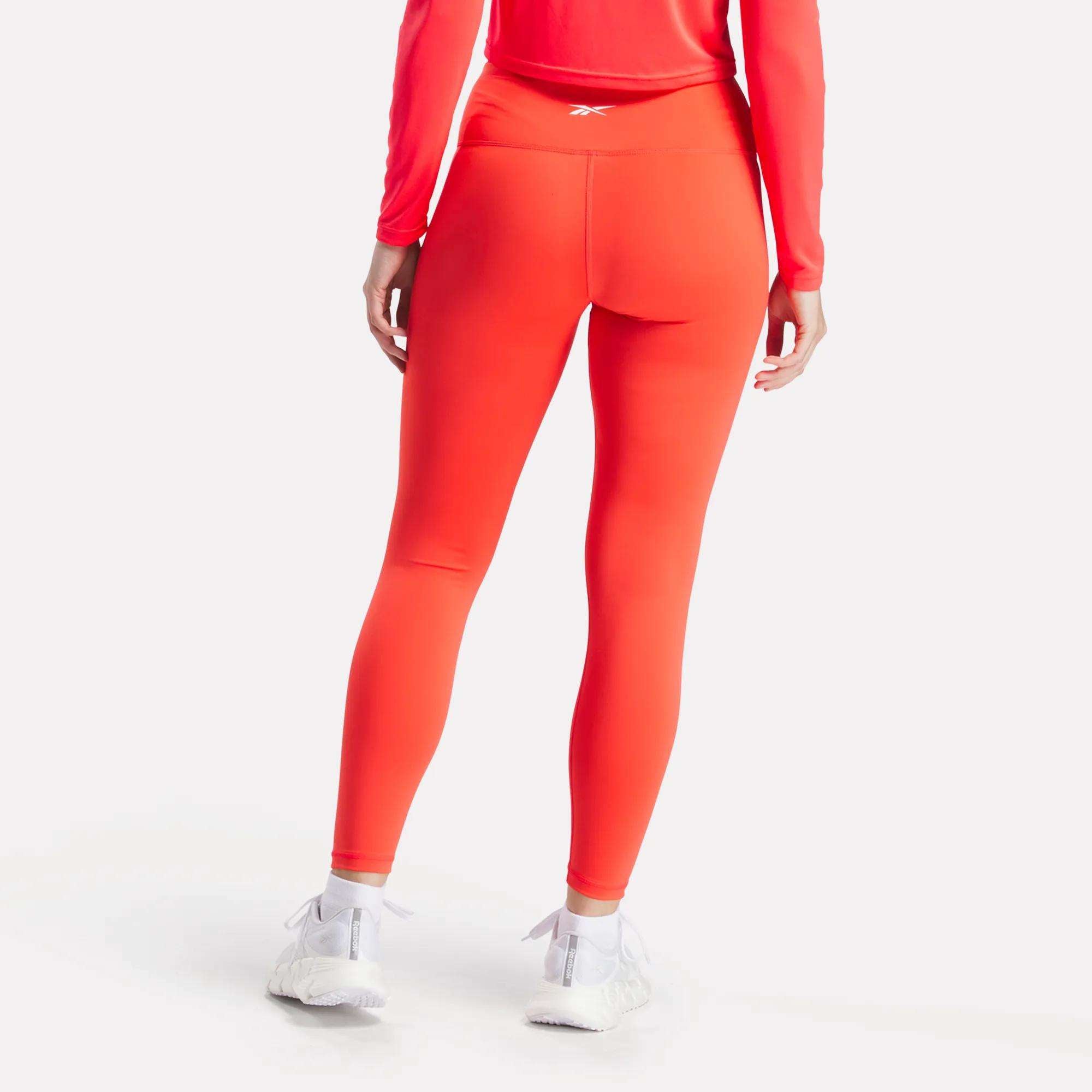 A person faces away, wearing Reebok Women's Core High-Rise Leggings in bright orange with a matching long-sleeve top and white athletic shoes against a plain white background.
