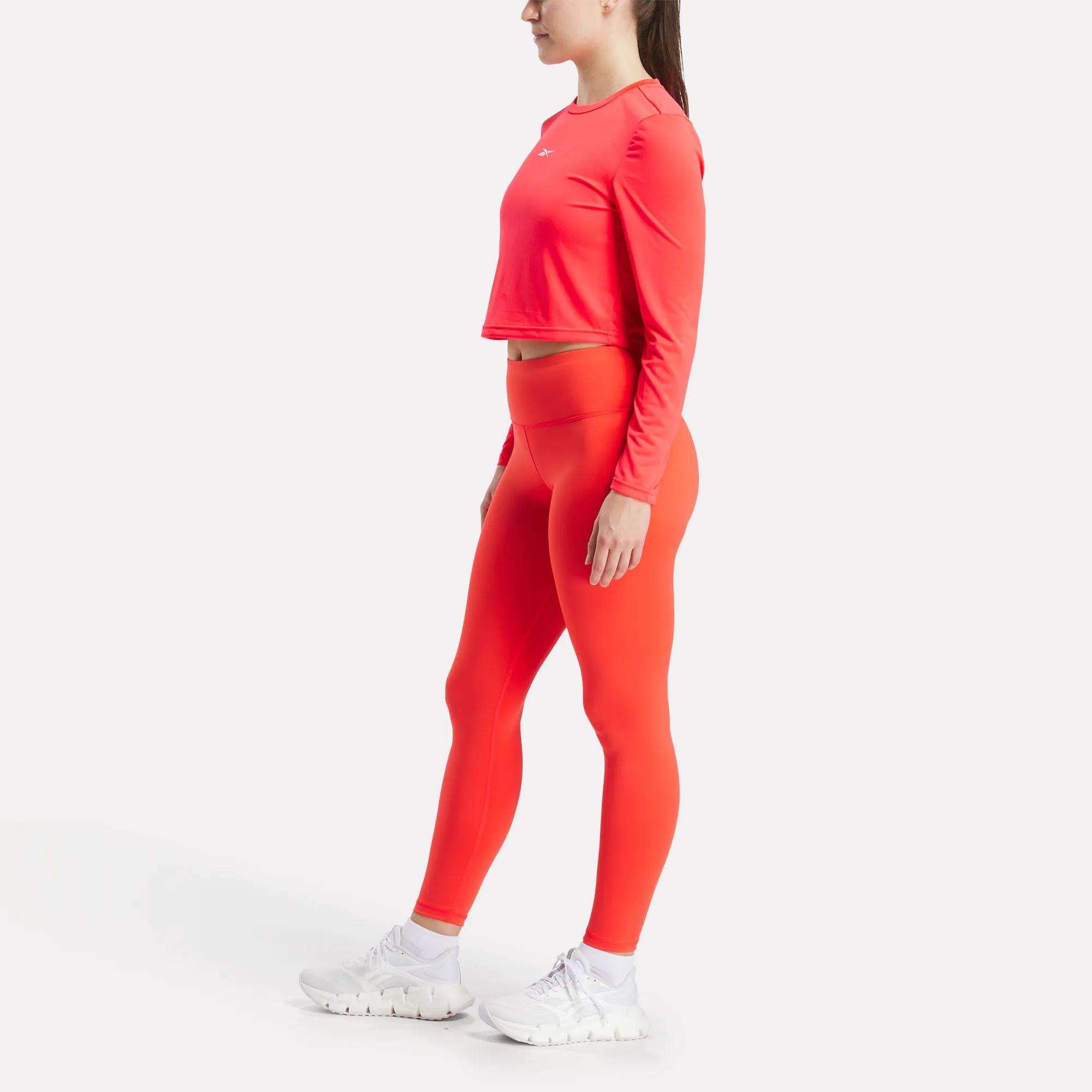 A woman stands sideways in a bright red long-sleeve crop top and Reebok Women's Core High-Rise Leggings, paired with white athletic shoes, against a plain white background.