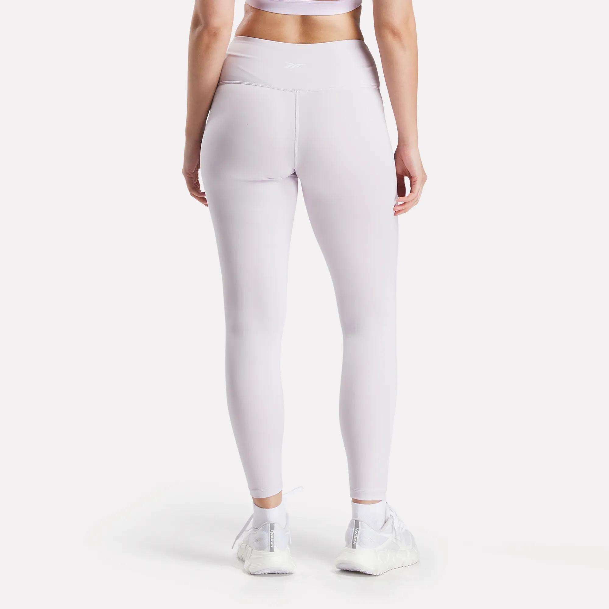 A person stands with their back to the camera against a white background, wearing matching light purple Reebok Women’s Core High-Rise Leggings and a sports bra, paired with white sneakers for a coordinated sporty look.